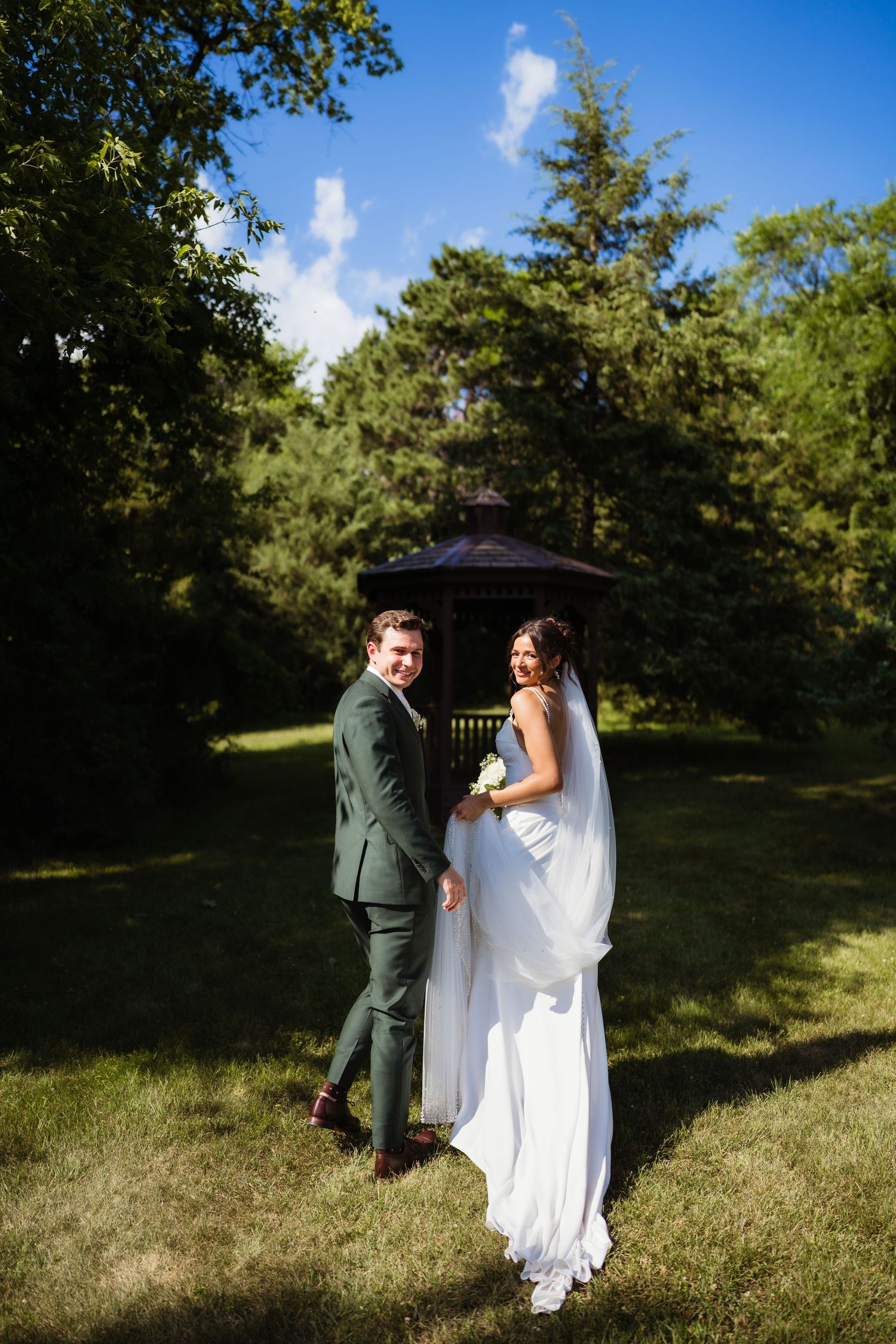 A bride and groom hold hands smiling in a lush green garden with a gazebo backdrop on their wedding day.