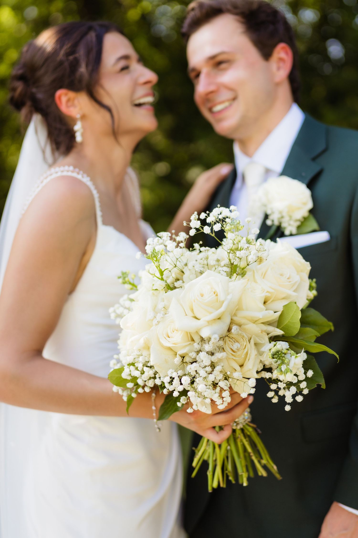 Smiling bride holding white rose bouquet with baby's breath stands with groom in green suit at outdoor wedding.