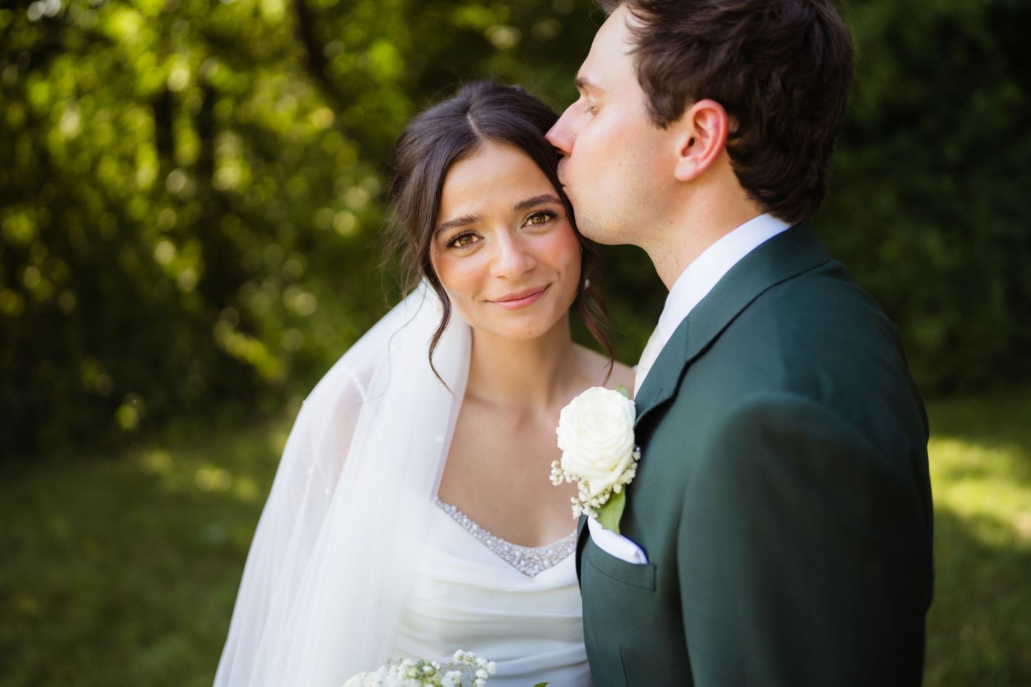 Groom in green suit kissing bride's cheek outdoors, bride wearing white gown and veil with floral bouquet.