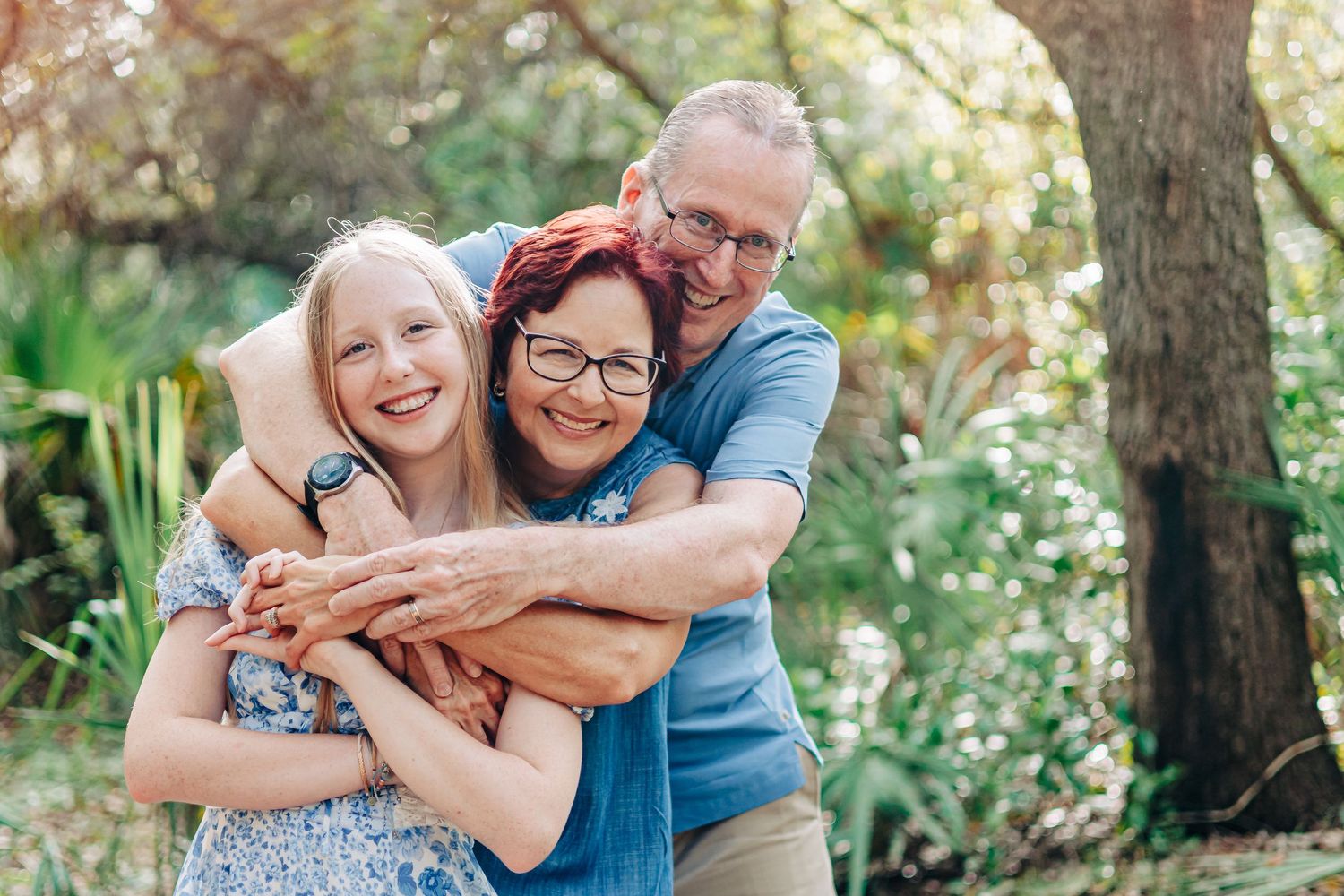 A joyful family of three sharing a warm hug and laughter outdoors in a sunlit garden setting at at Delray Oaks Nature trail Boca Raton FL..
