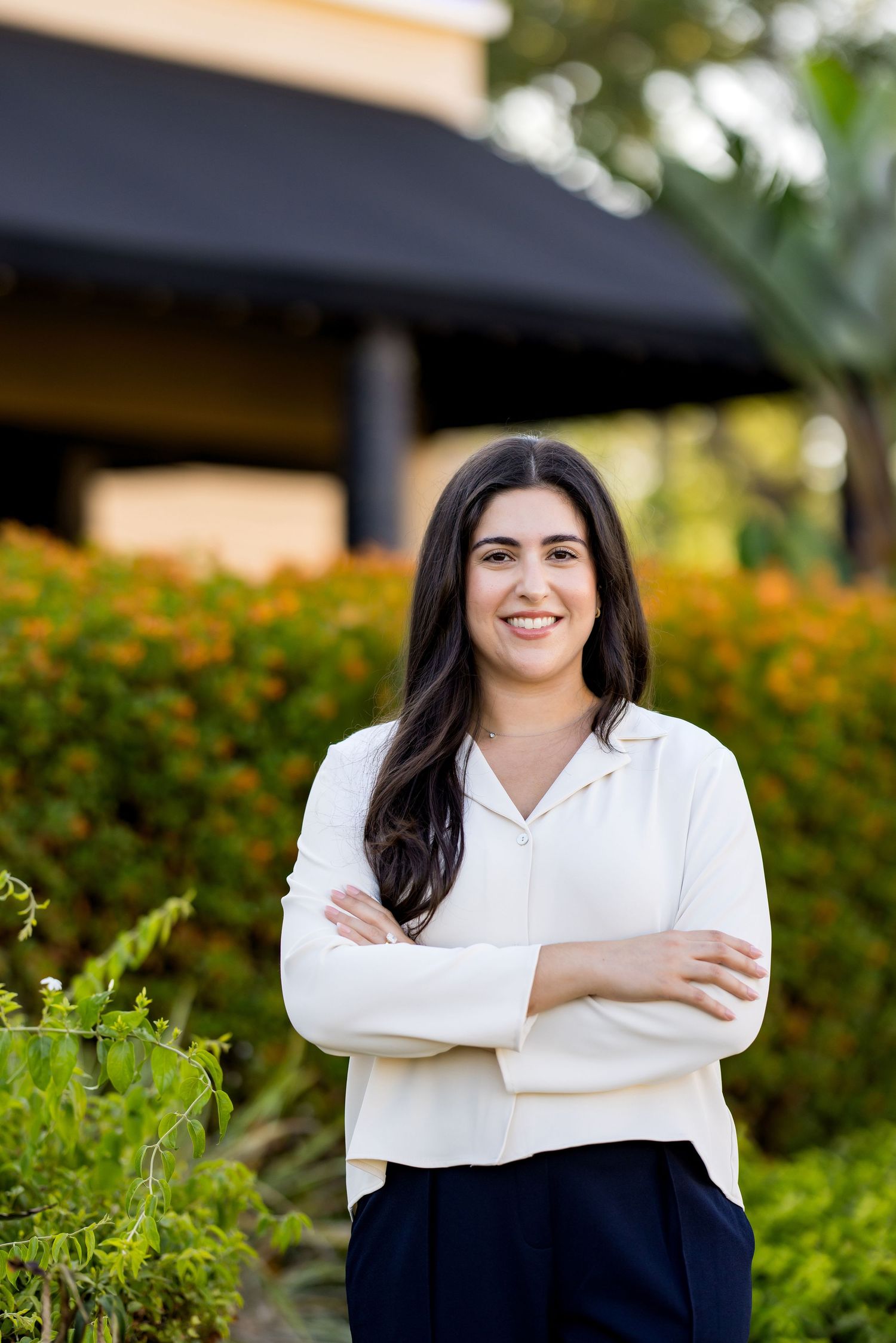 Professional woman in white blouse stands confidently with arms crossed, smiling outdoors near colorful garden.