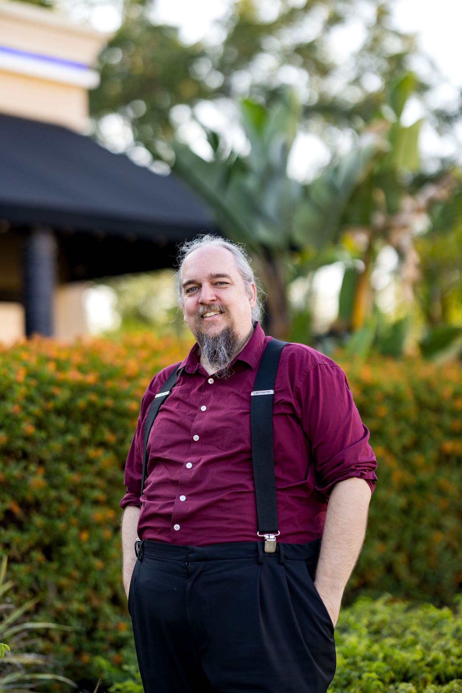Smiling middle-aged man with gray beard wearing maroon shirt and suspenders standing outdoors near tropical greenery.