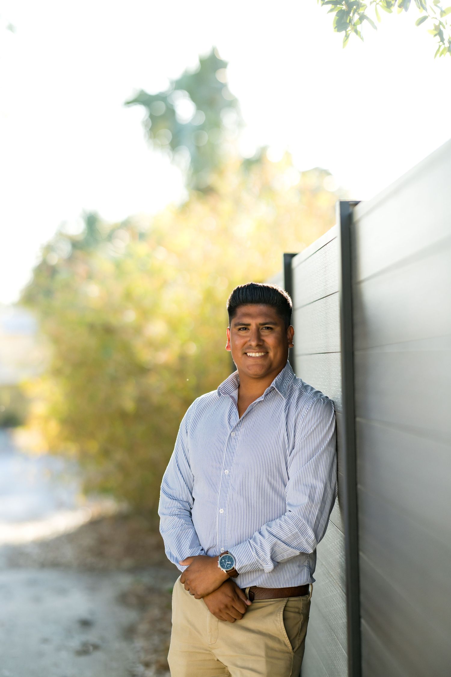 Smiling man in light blue shirt and khaki pants leaning against gray wall outdoors with autumn trees.