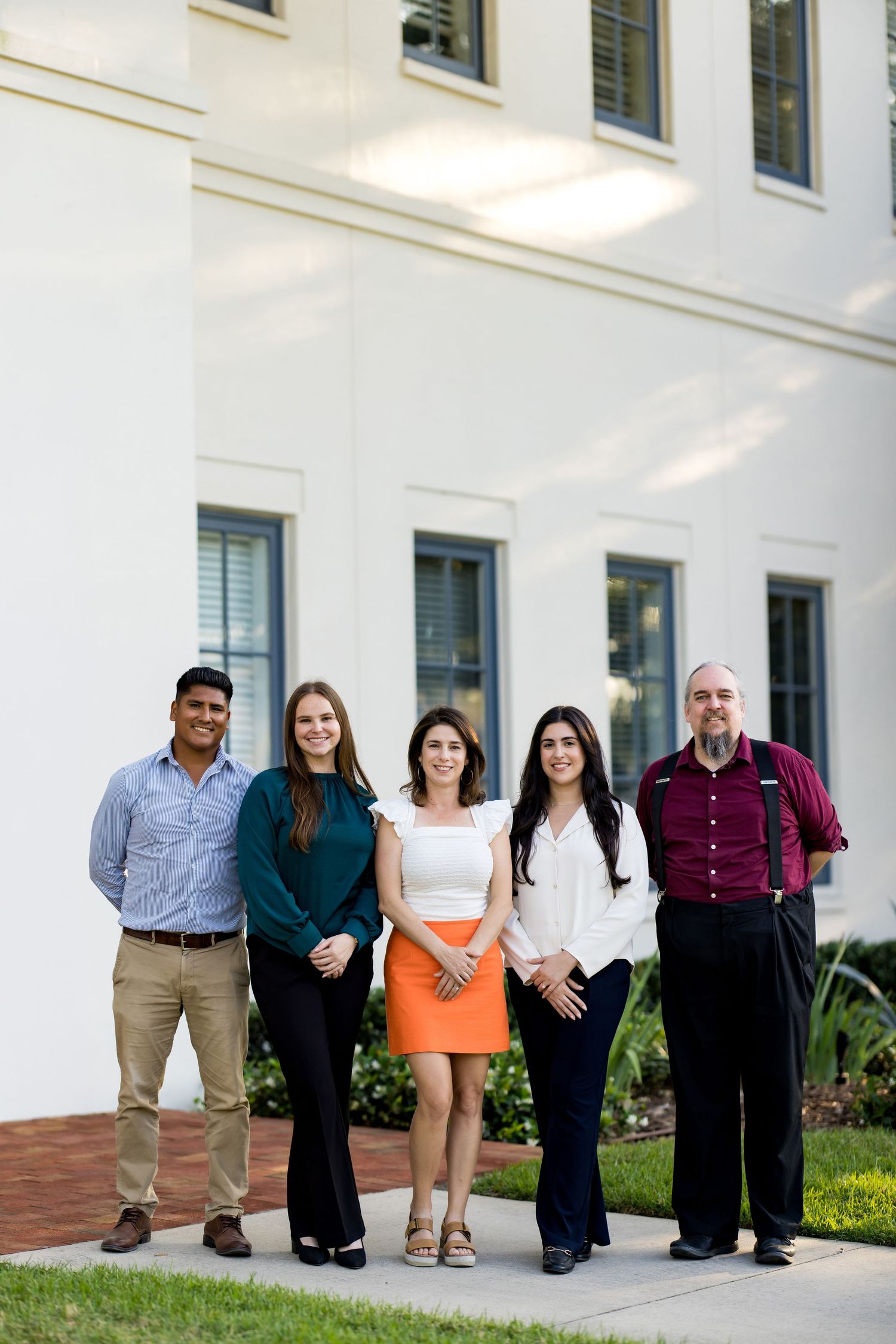 Five professionals pose together outdoors in front of a white building, dressed in business casual attire.