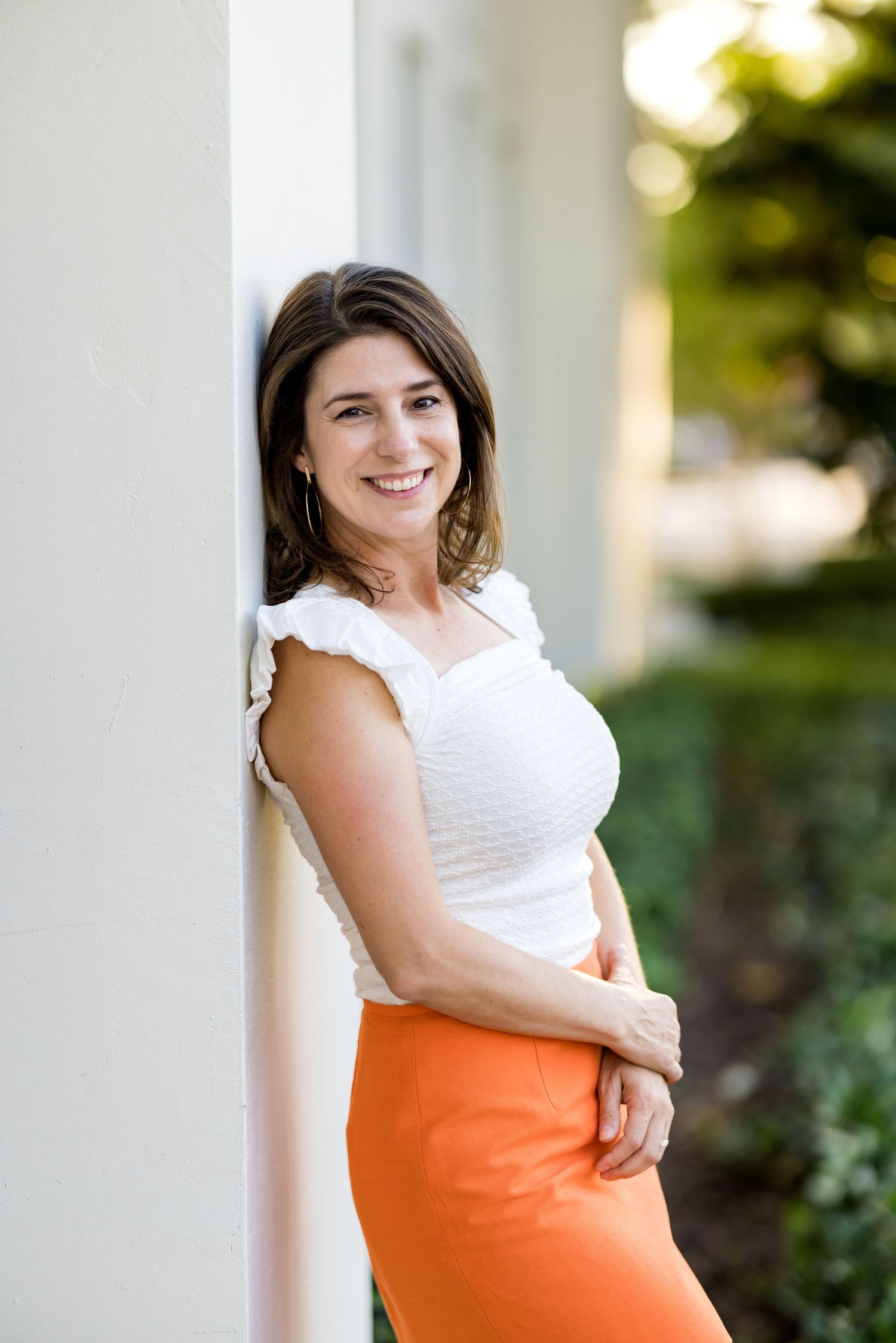 Smiling woman in white top and orange skirt leaning against a white pillar outdoors.
