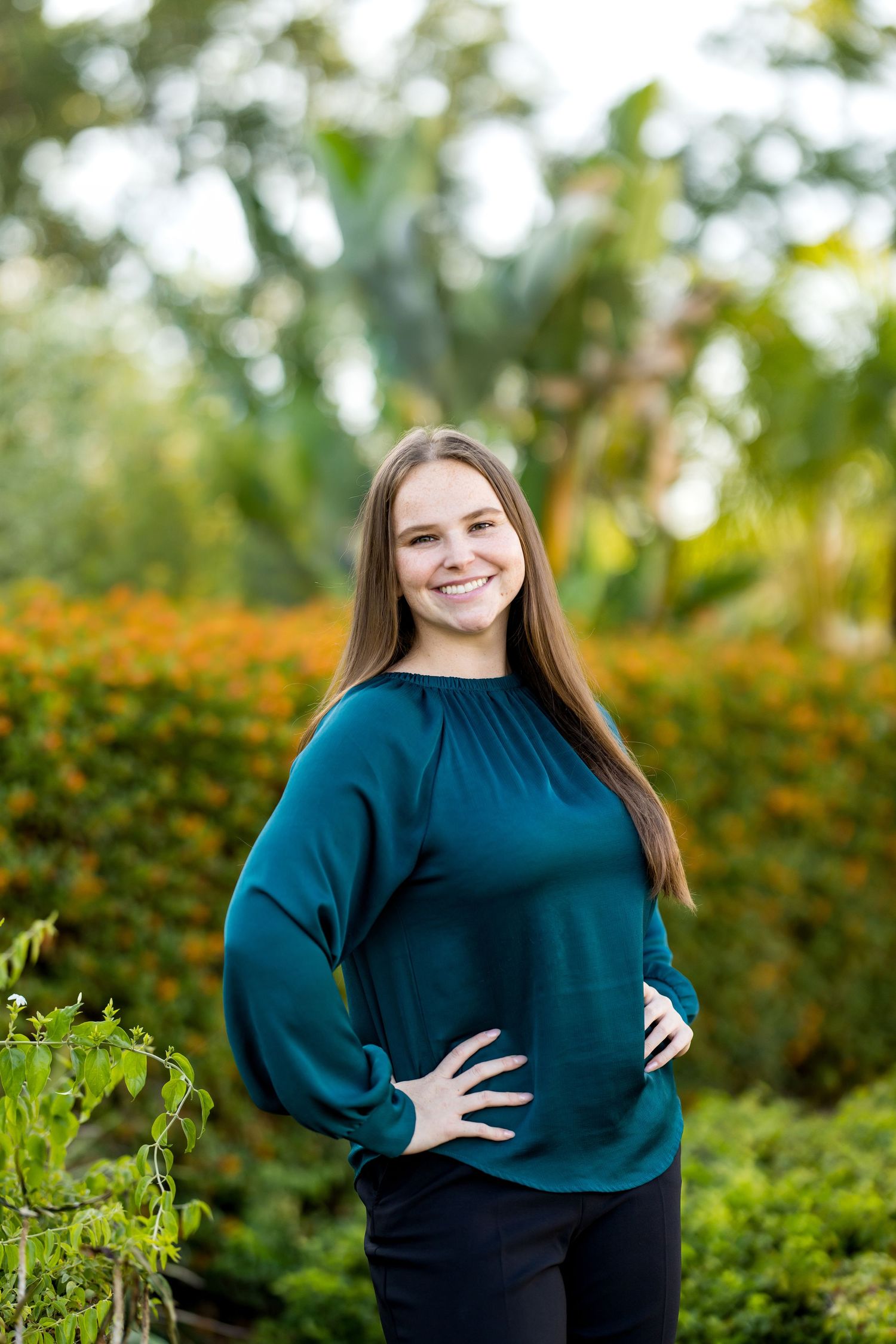 Young woman in teal blouse smiling outdoors with lush green garden and orange flowers in background.