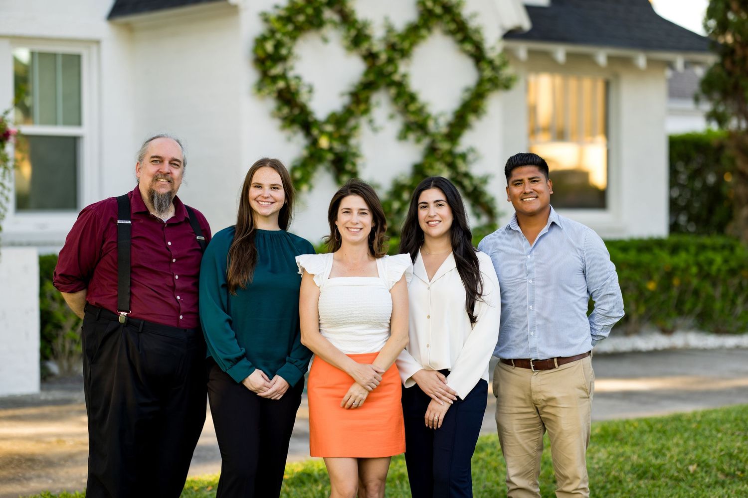Five smiling family members posing together outdoors in front of a white house with festive wreath decorations.