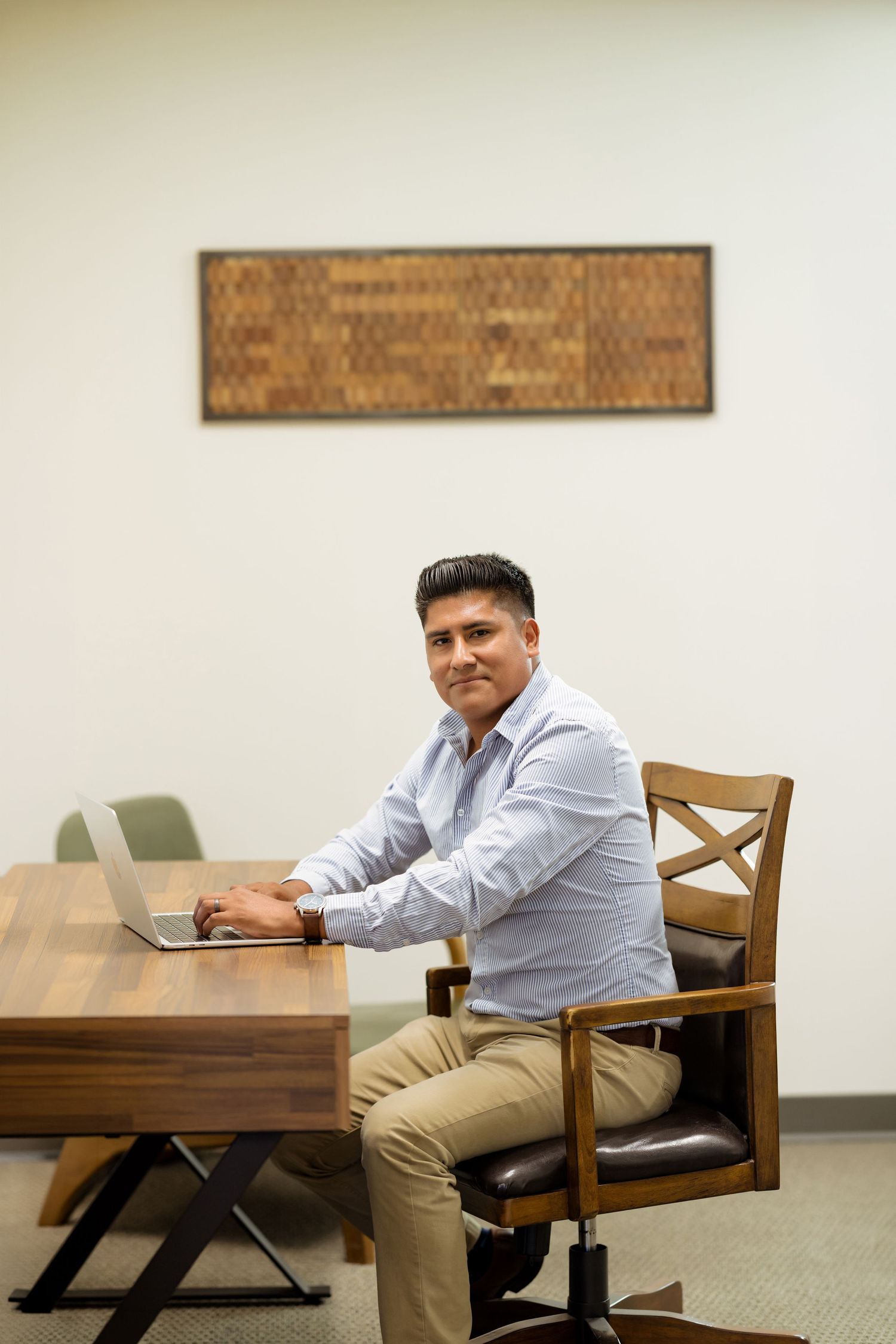 A professional man in a light blue shirt sits at a wooden desk with a laptop in a modern office setting.
