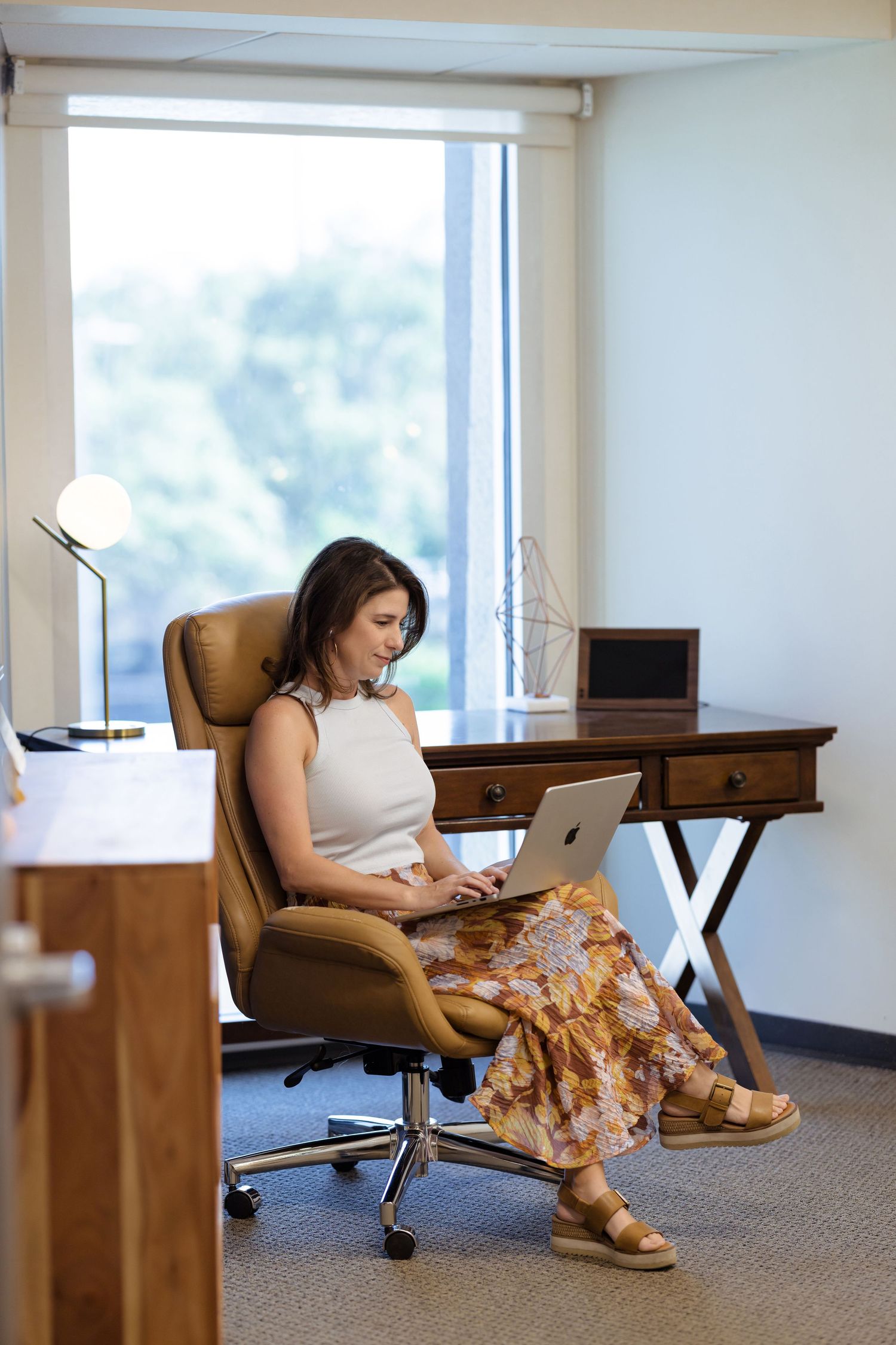Woman in floral skirt working on laptop in modern office chair at wooden desk near window.