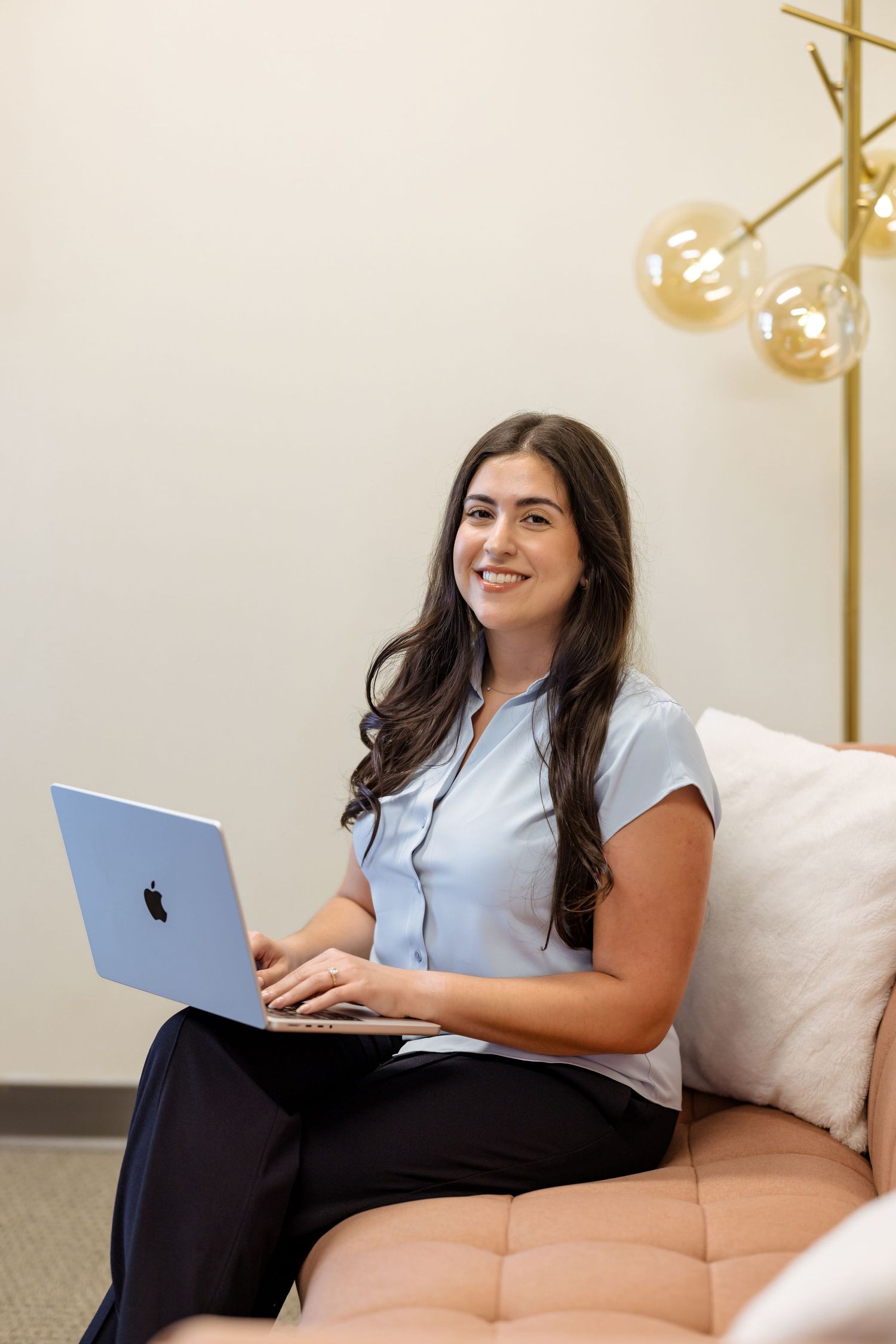 Young professional woman smiling with blue MacBook laptop, seated on couch in modern office setting.