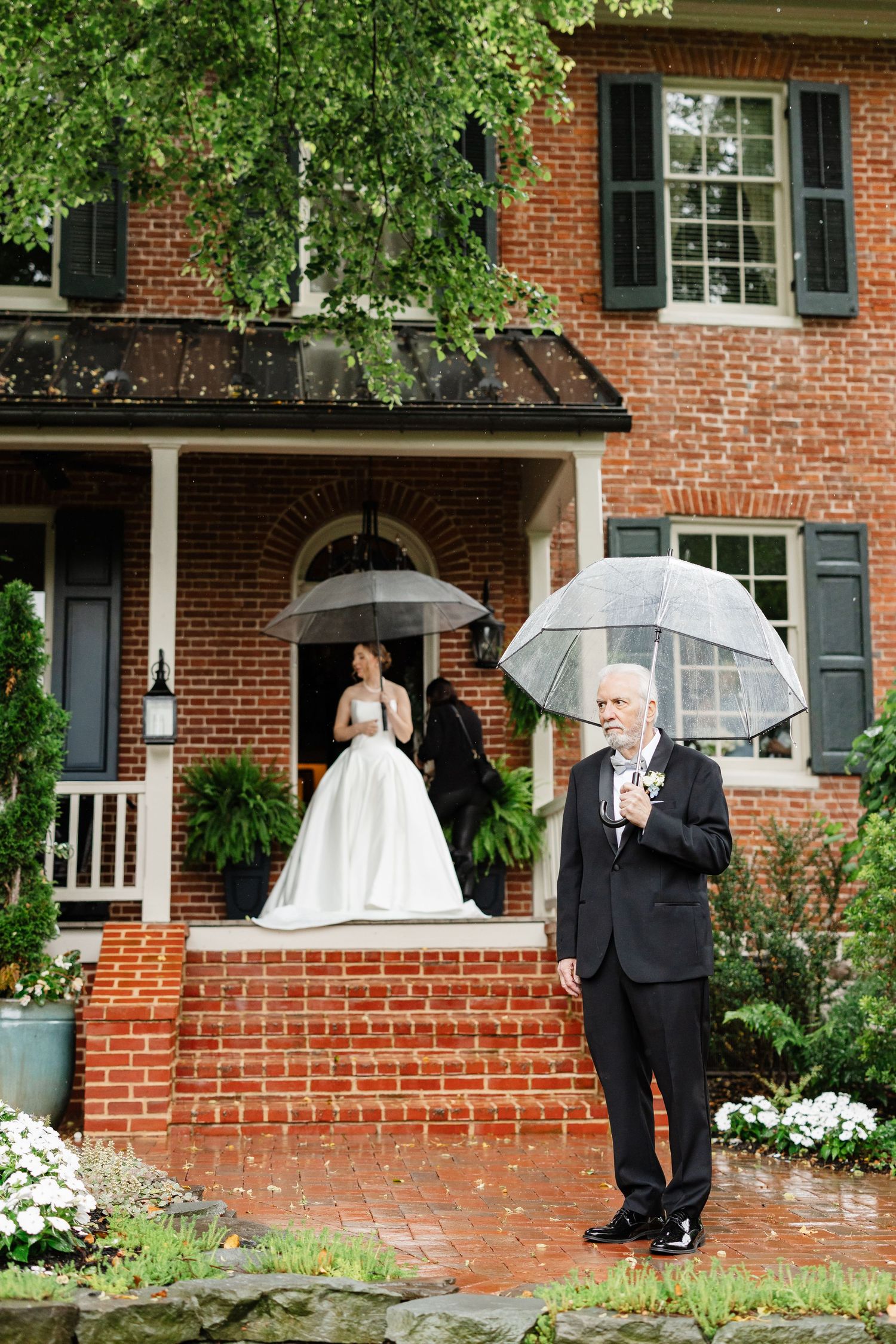 Bride in white ball gown stands on brick steps of red brick manor as escort waits with umbrella.