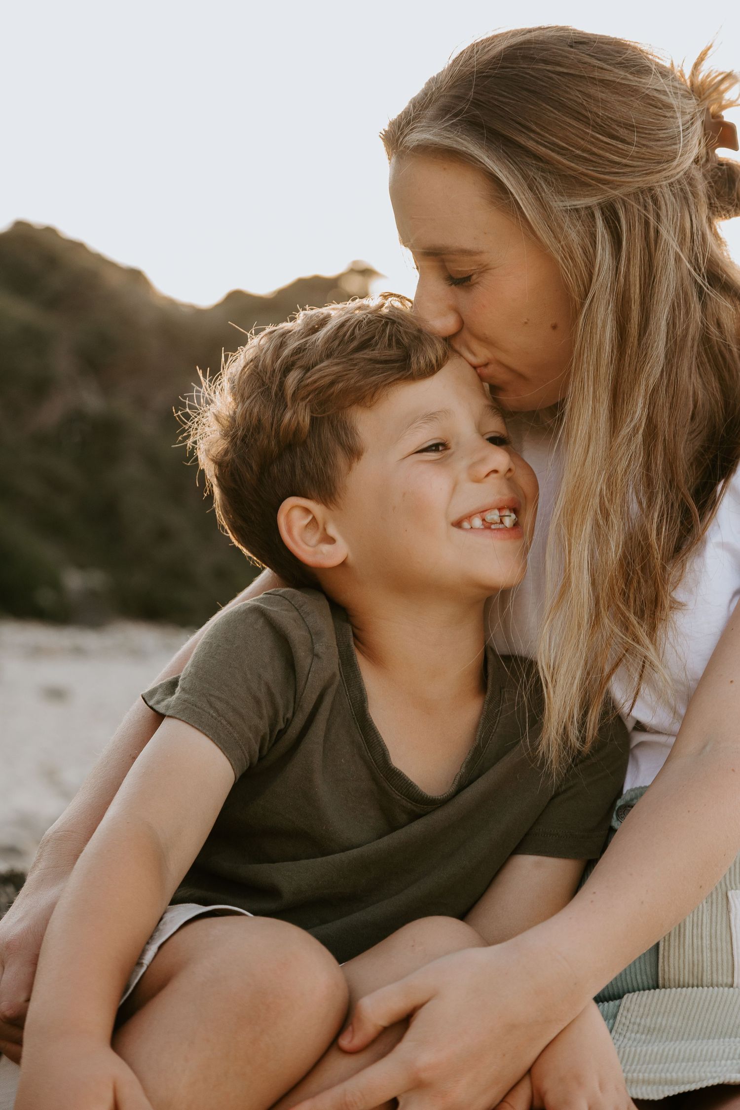 Family of four sitting on rocky beach at golden sunset with lush green hillside backdrop.