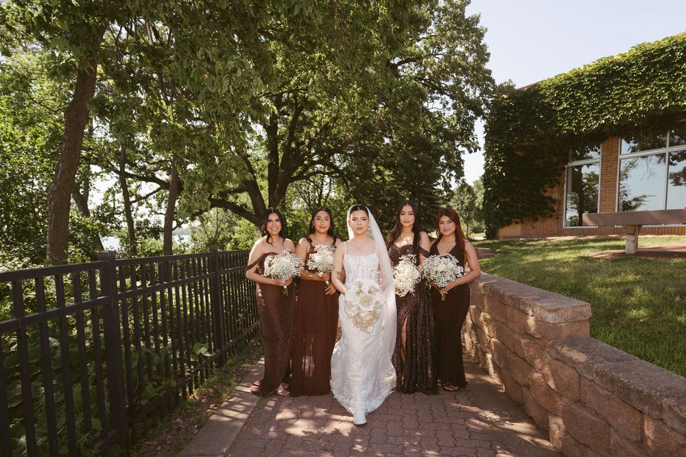 Bride in white gown walks with bridesmaids in brown dresses along a garden path surrounded by lush green trees.