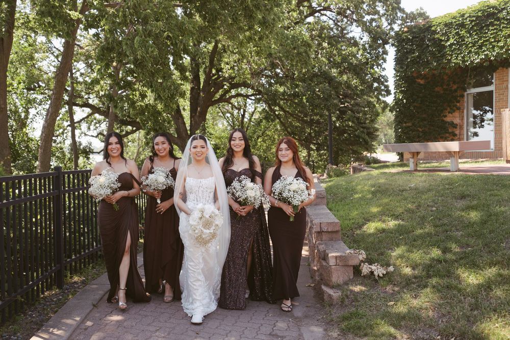 Bride in white lace gown walks with four bridesmaids in dark dresses through a tree-lined outdoor venue.