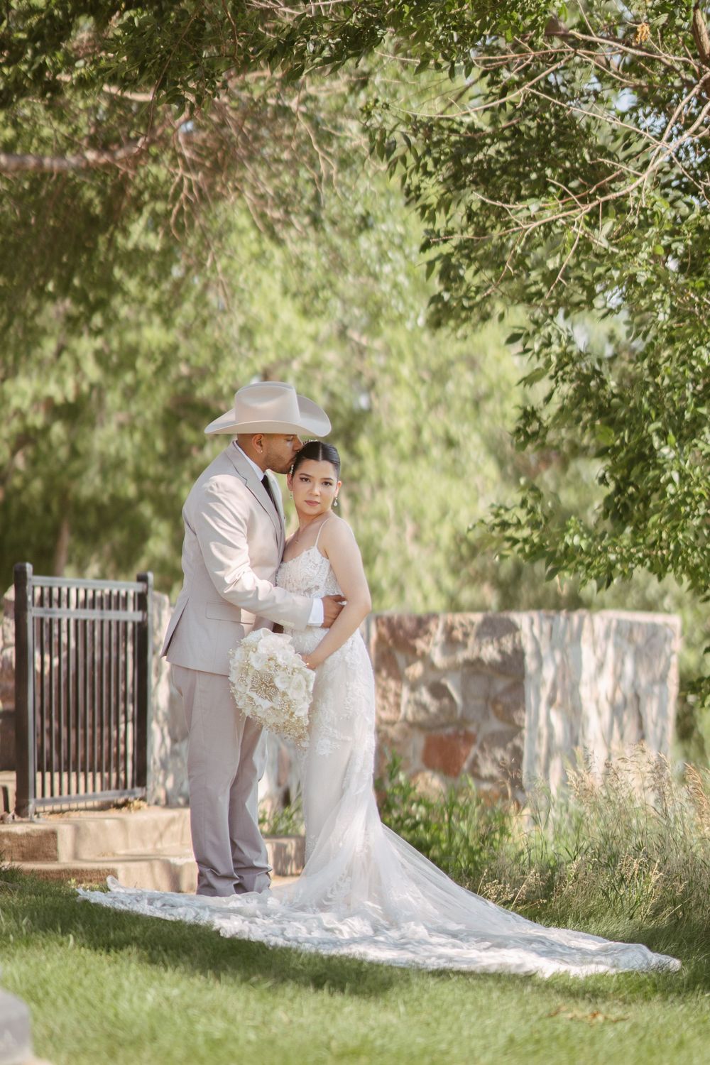 Bride in white lace gown holds bouquet as groom in tan suit and cowboy hat embraces her in lush garden.