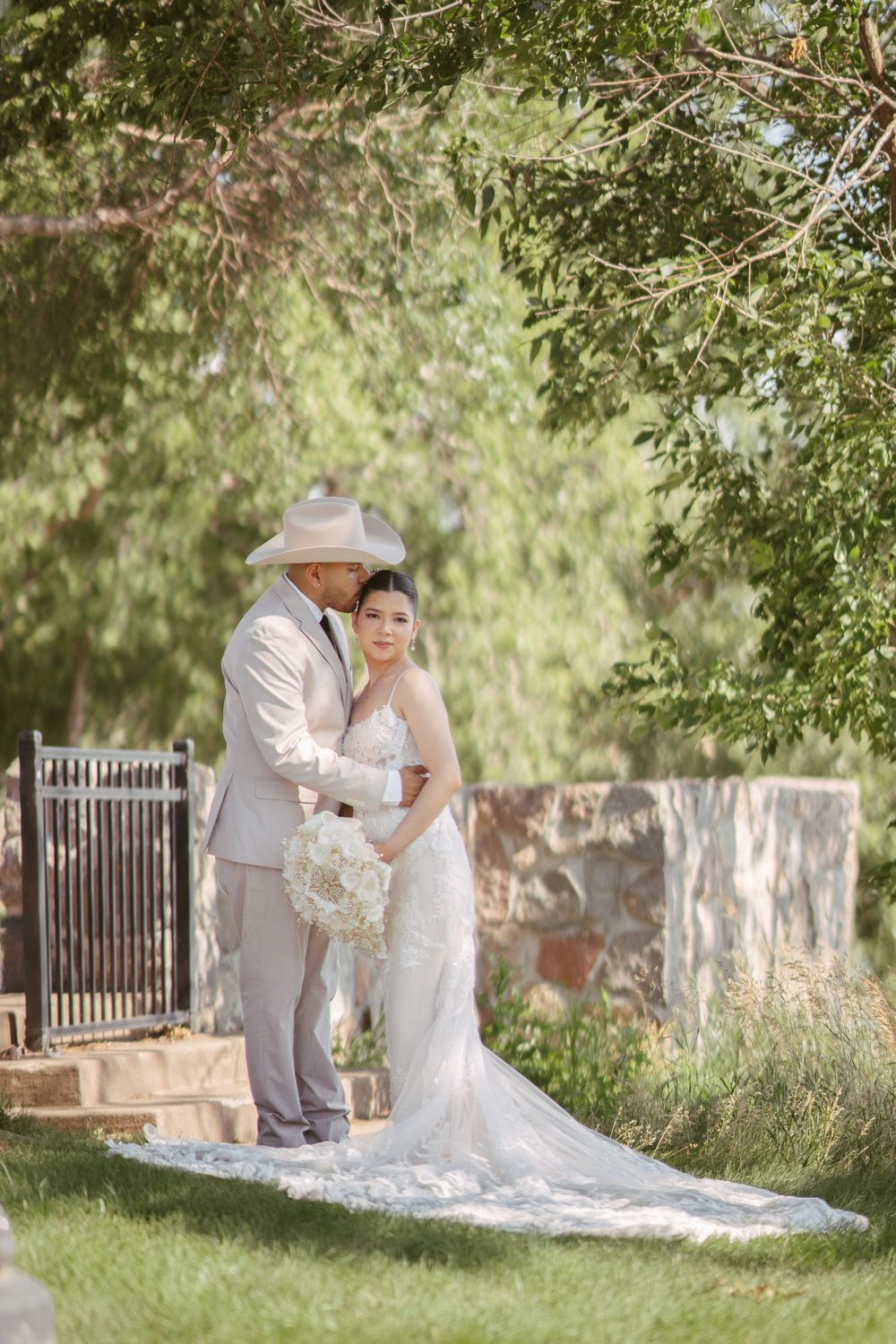 Bride in white gown with groom in tan suit and cowboy hat embracing outdoors under green trees.