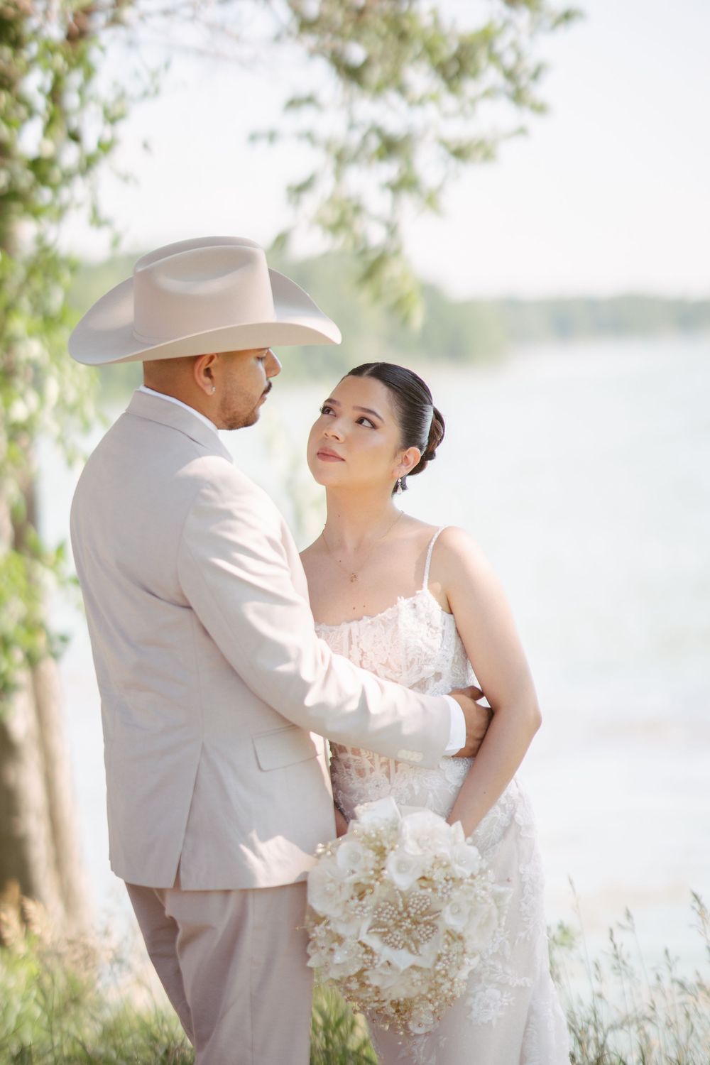 Bride and groom share a romantic moment by a lakeside, groom wearing a white cowboy hat and tan suit, bride holding white bouquet.