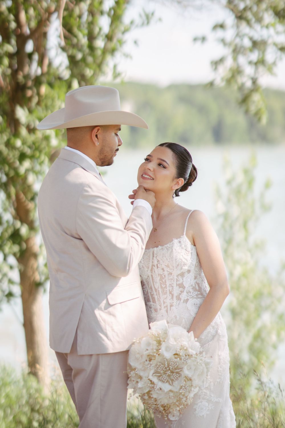 A groom in a beige suit and cowboy hat tenderly holds his bride's face as she smiles, holding a white bouquet outdoors.