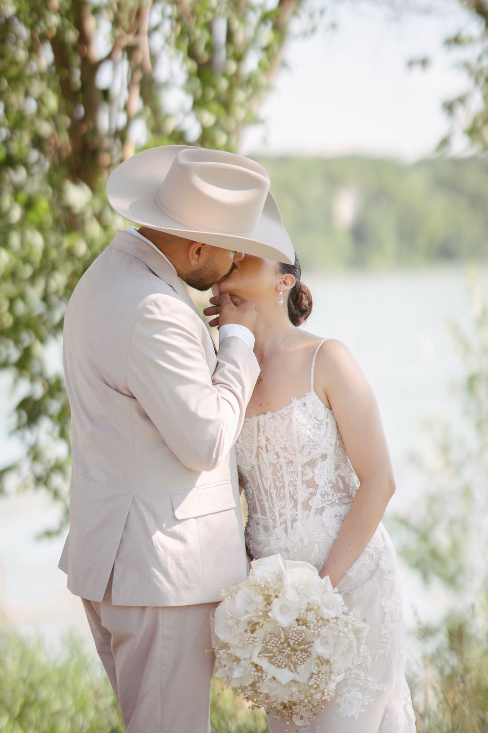 Bride and groom share a romantic kiss outdoors near a lake, groom wearing a beige suit and cowboy hat.