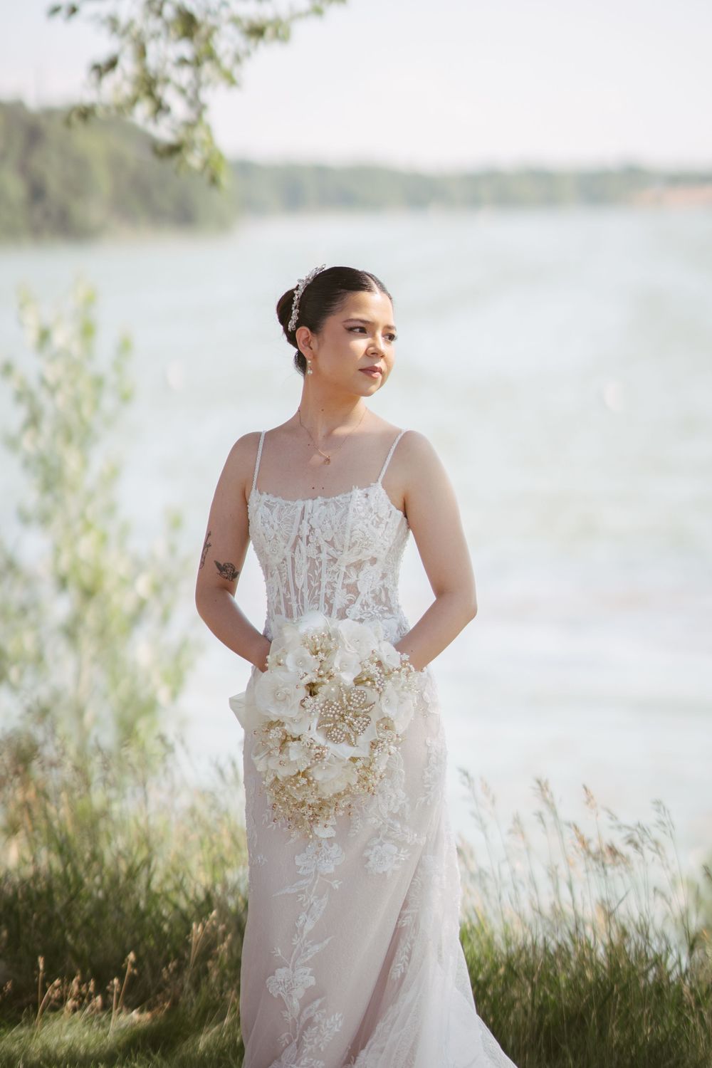 Bride in lace wedding gown holding white bouquet by lakeside with greenery backdrop.