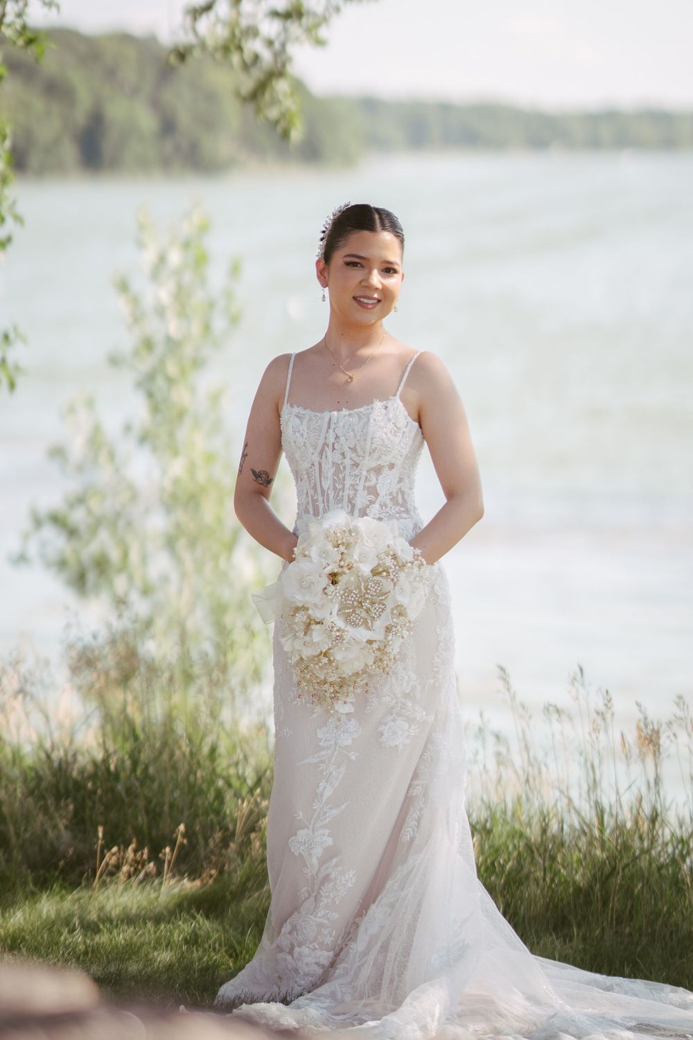 Bride in lace wedding gown holding white bouquet by a lakeside.