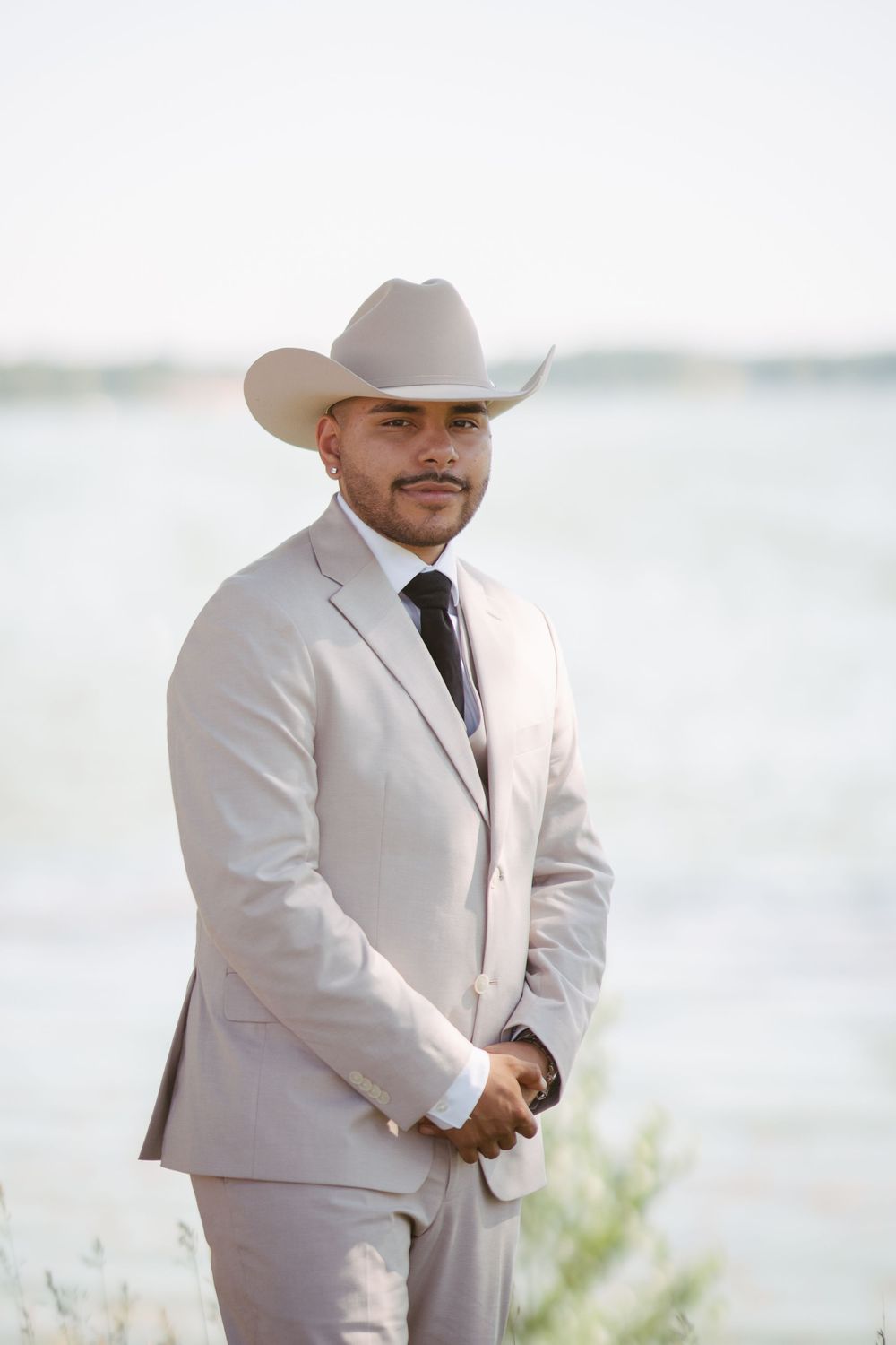 Groom in light gray suit and cowboy hat poses confidently near waterfront on wedding day.