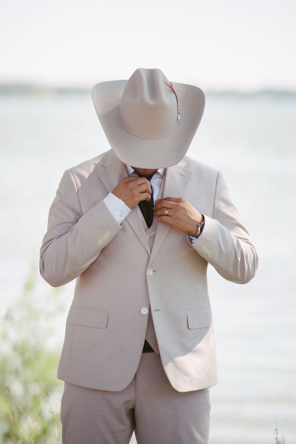 Groom in beige suit and cowboy hat adjusts his tie near a lakeside outdoor wedding venue.