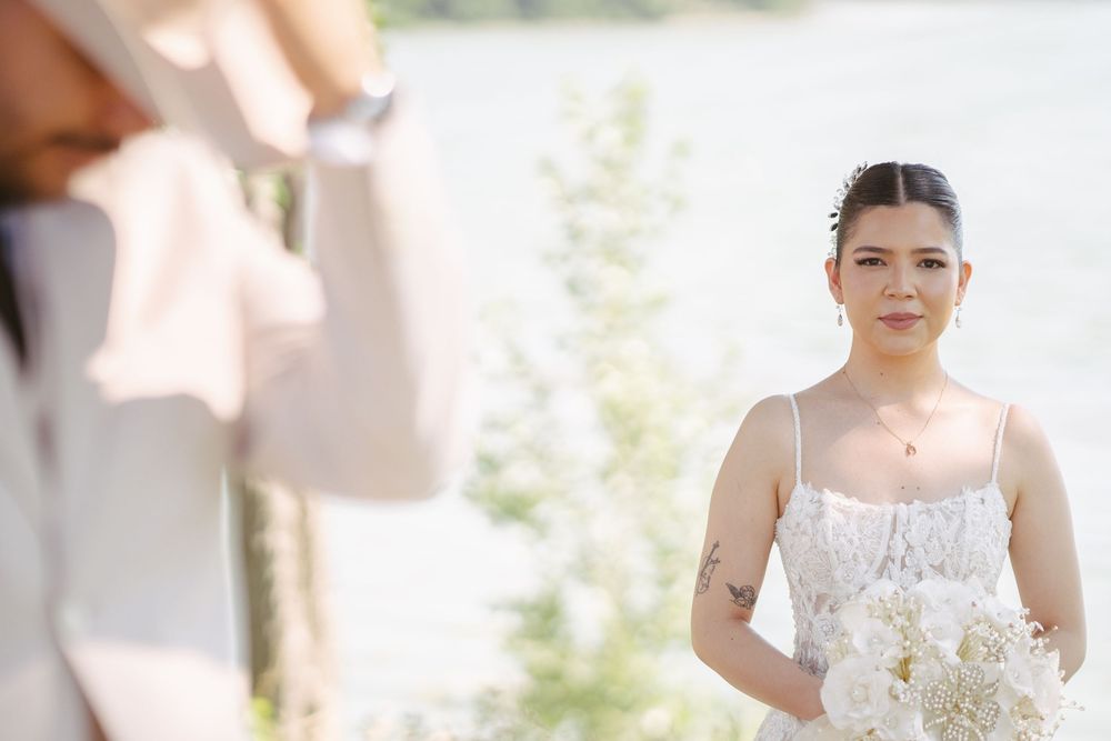 Bride in white dress holding bouquet of white flowers outdoors near water, being photographed at wedding ceremony.