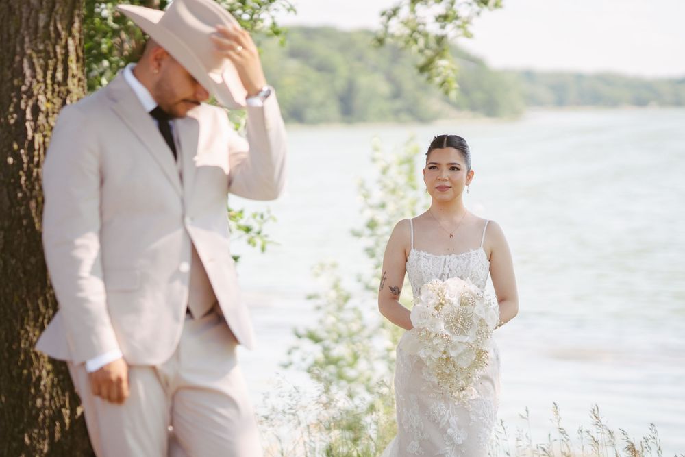 Bride in lace gown holding white bouquet approaches groom in cream suit near lakeside trees at outdoor wedding.