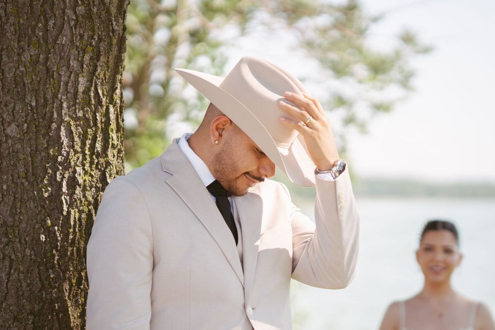 Groom in white suit and cowboy hat adjusting brim outdoors near tree with bride blurred in background.