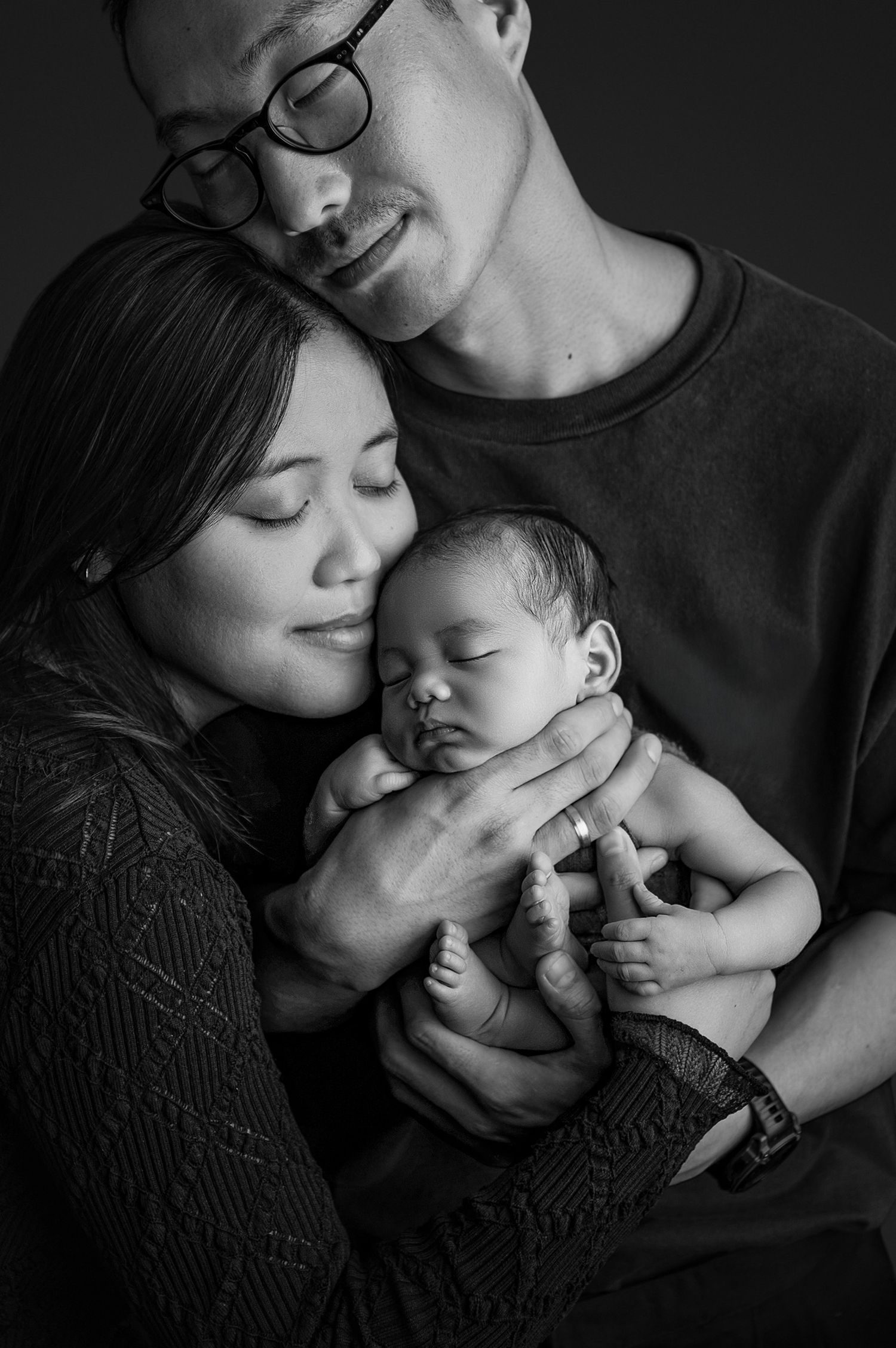 Black and white portrait of Asian parents tenderly cradling their sleeping newborn baby in a studio setting.