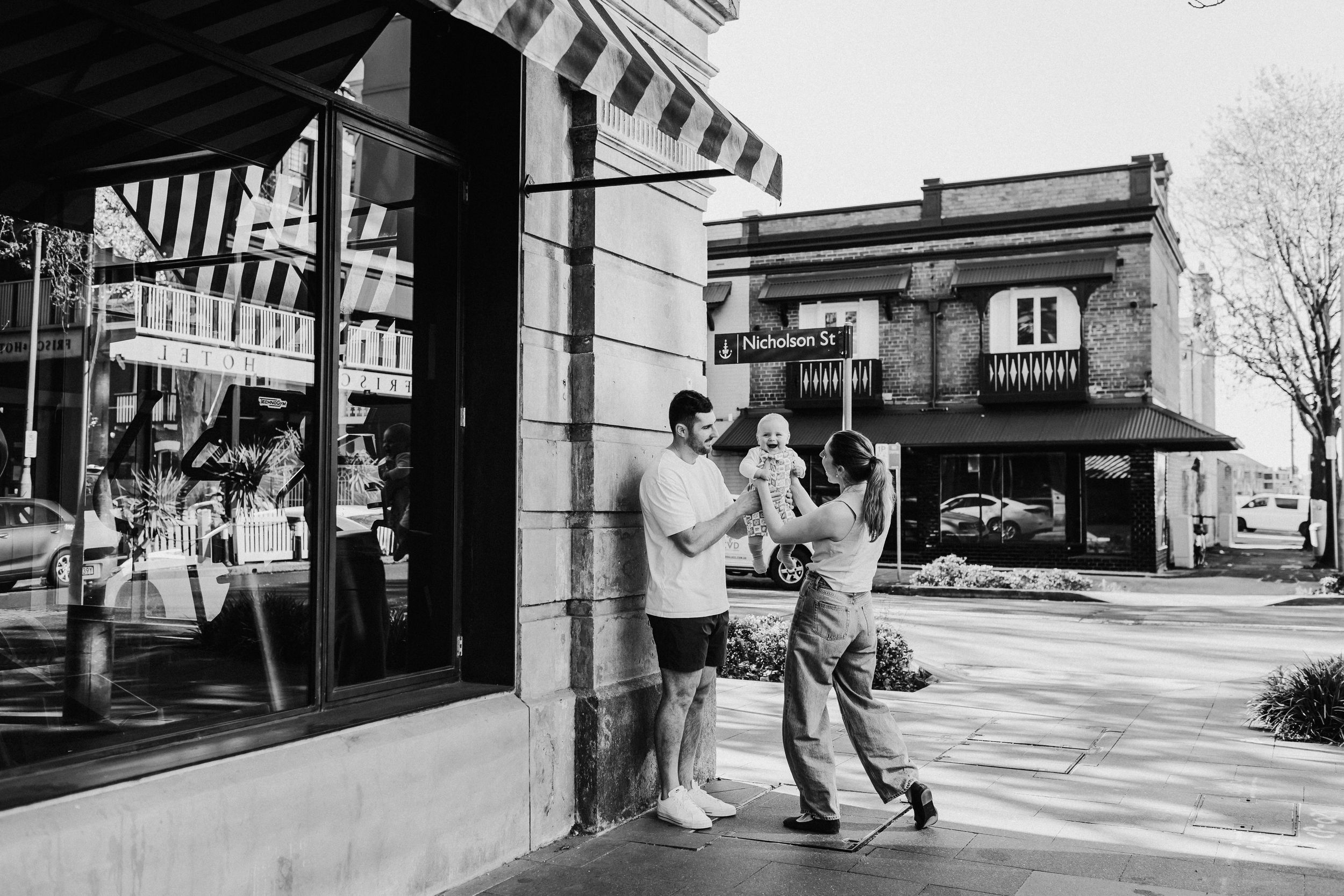 Black and white photo of a young family with baby on a charming small town sidewalk outside a shop.