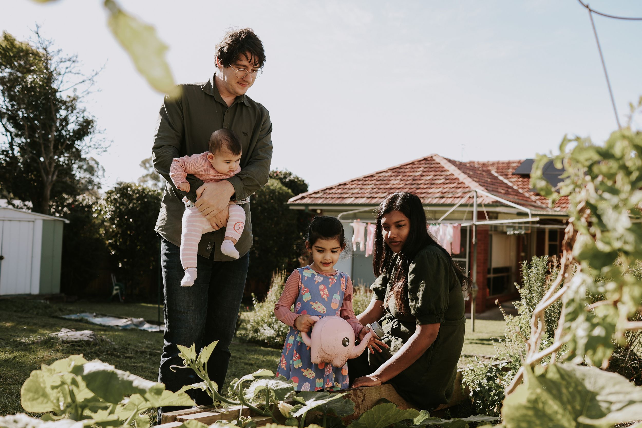 Family of four enjoying gardening together in backyard, father holding baby while girl waters plants near mother.