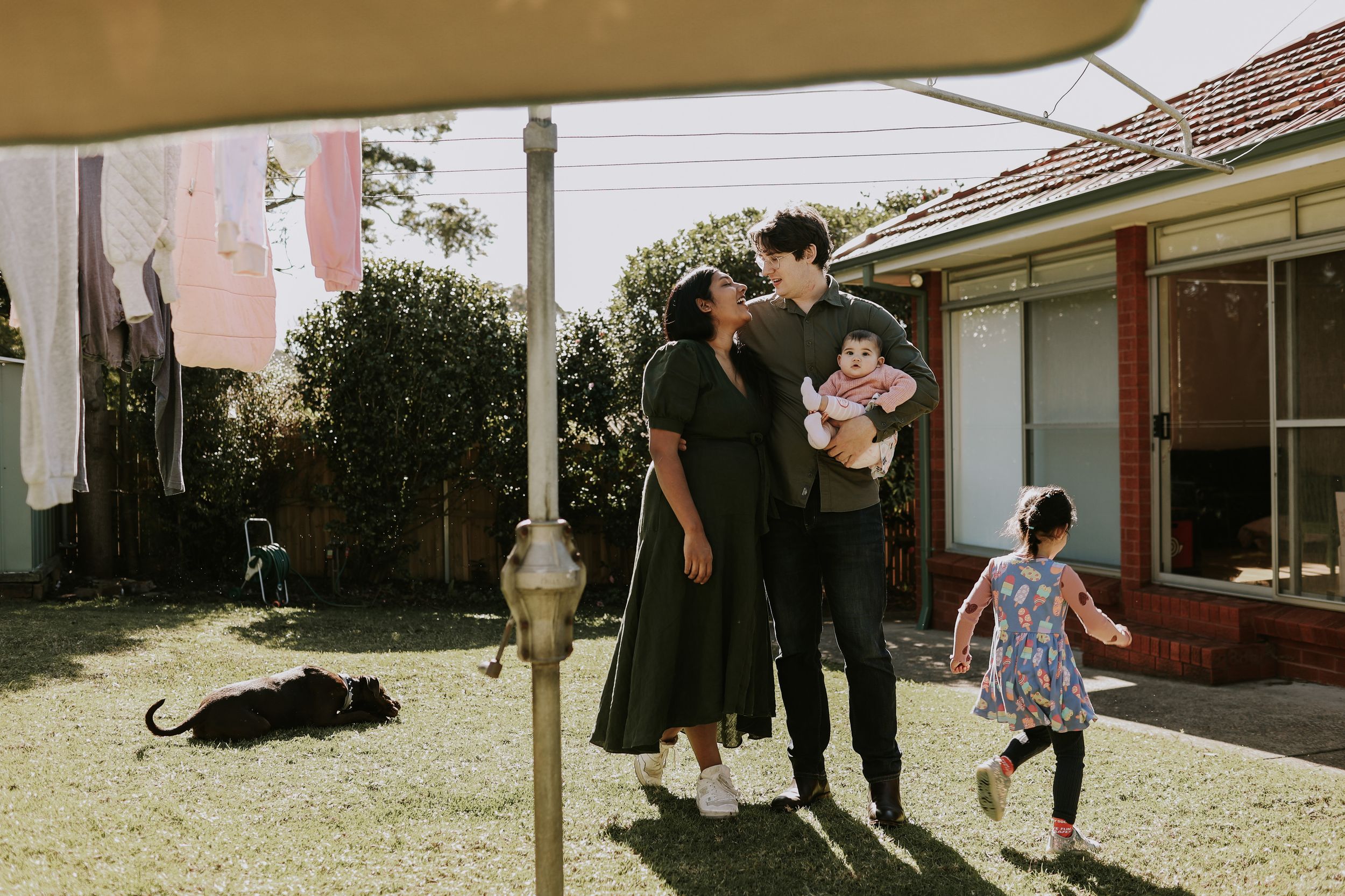 A young family stands in a sunny backyard near a clothesline, holding a baby while a child plays nearby.