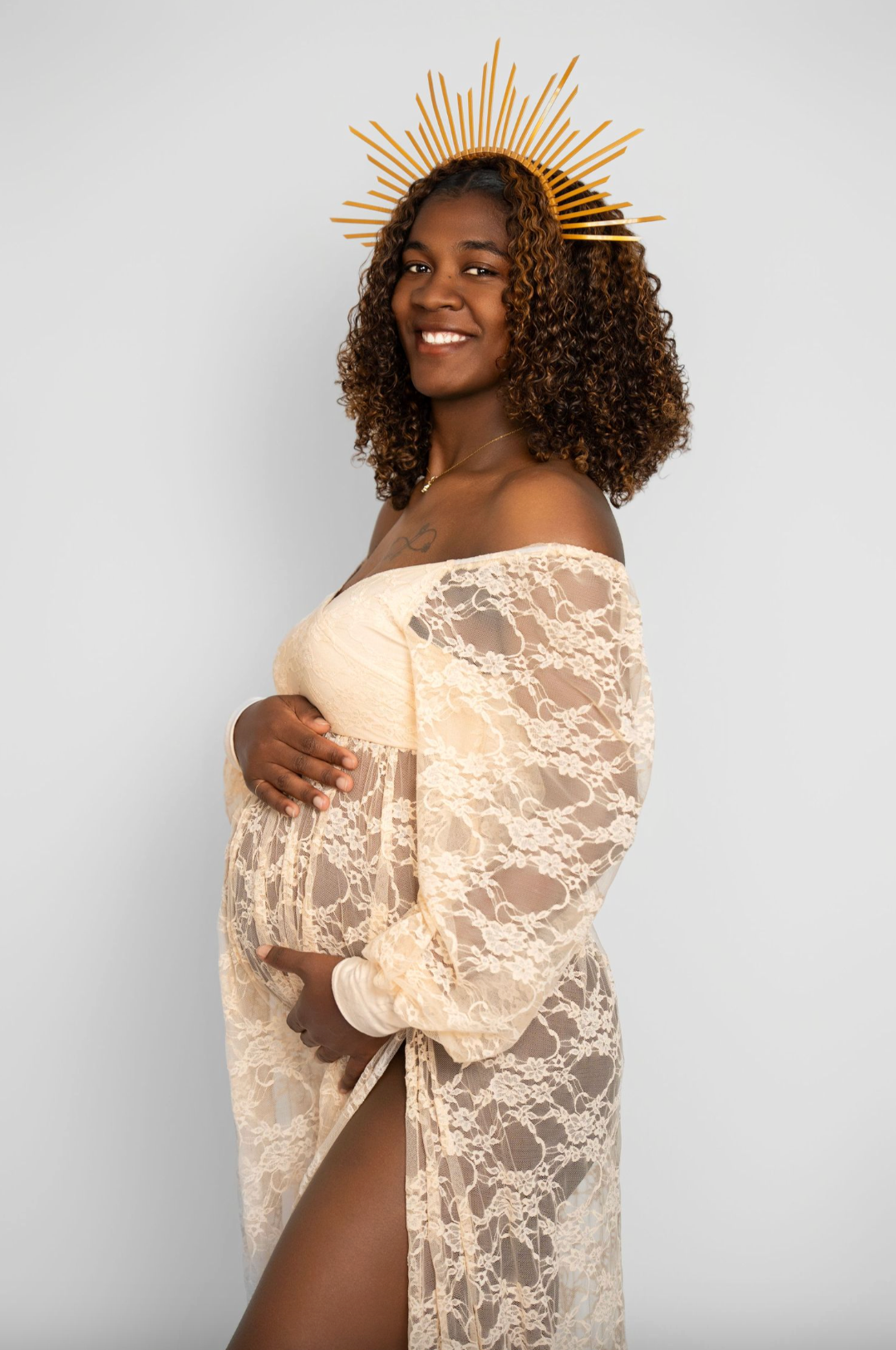 Smiling pregnant woman in sheer lace off-shoulder gown with gold sunburst crown, posing sideways against white background.