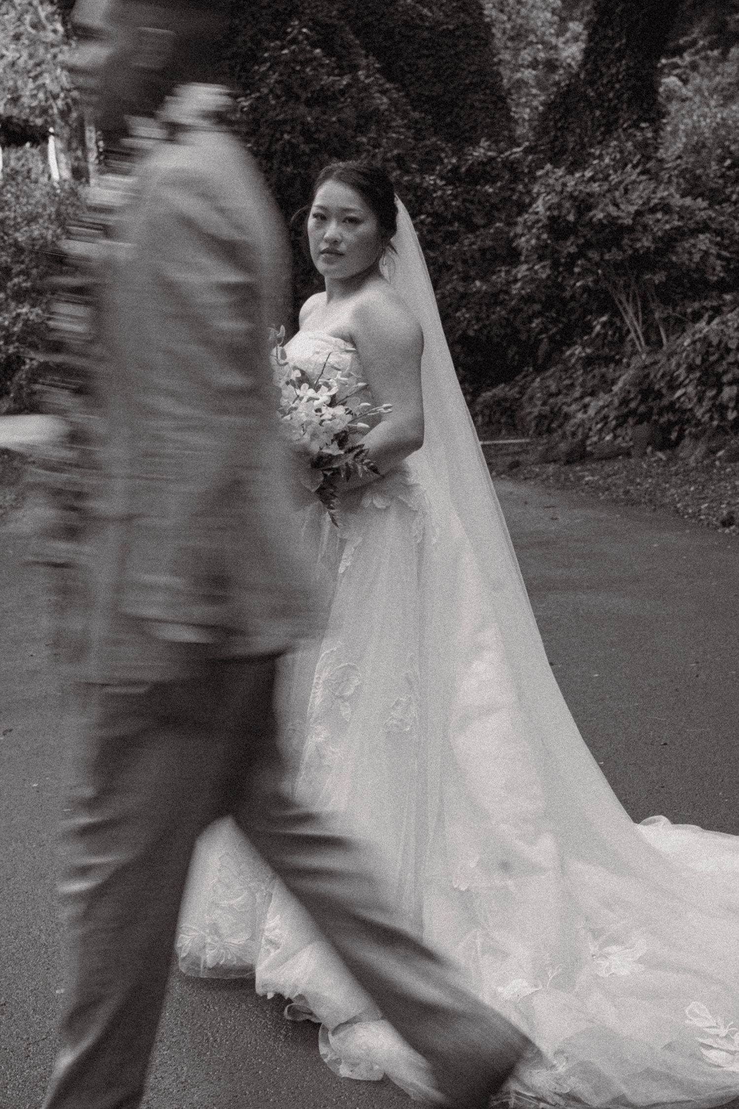 Black and white wedding photo of bride in lace gown holding bouquet, groom walking past in blurred motion.