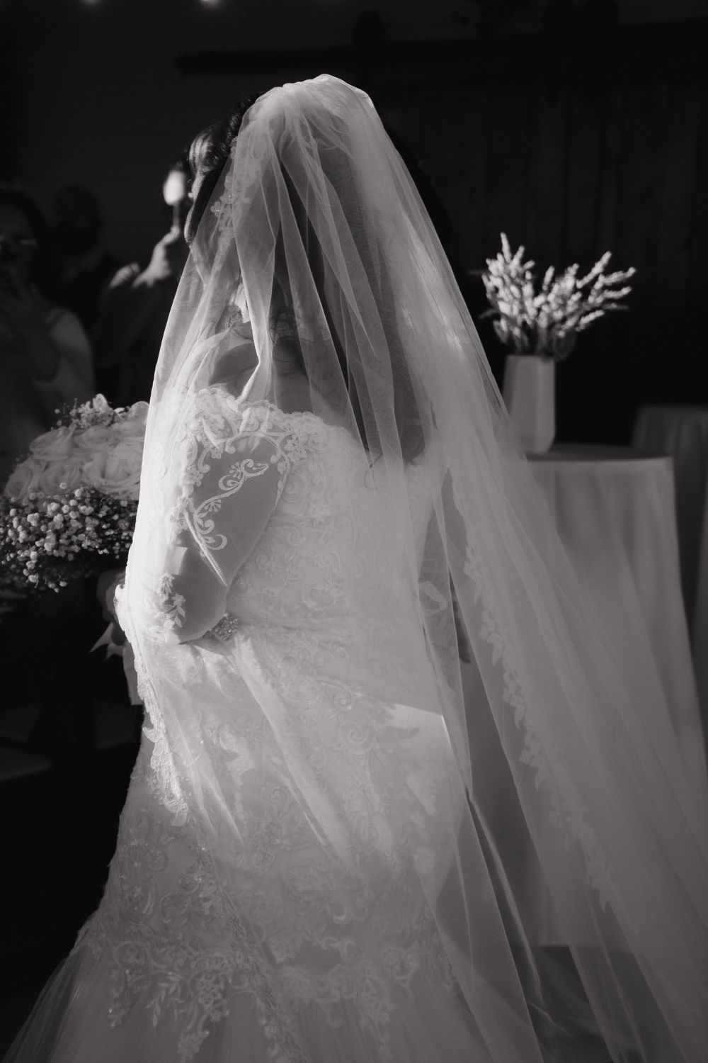 Black and white photo of a bride in a lace wedding dress and veil seen from behind at a ceremony.
