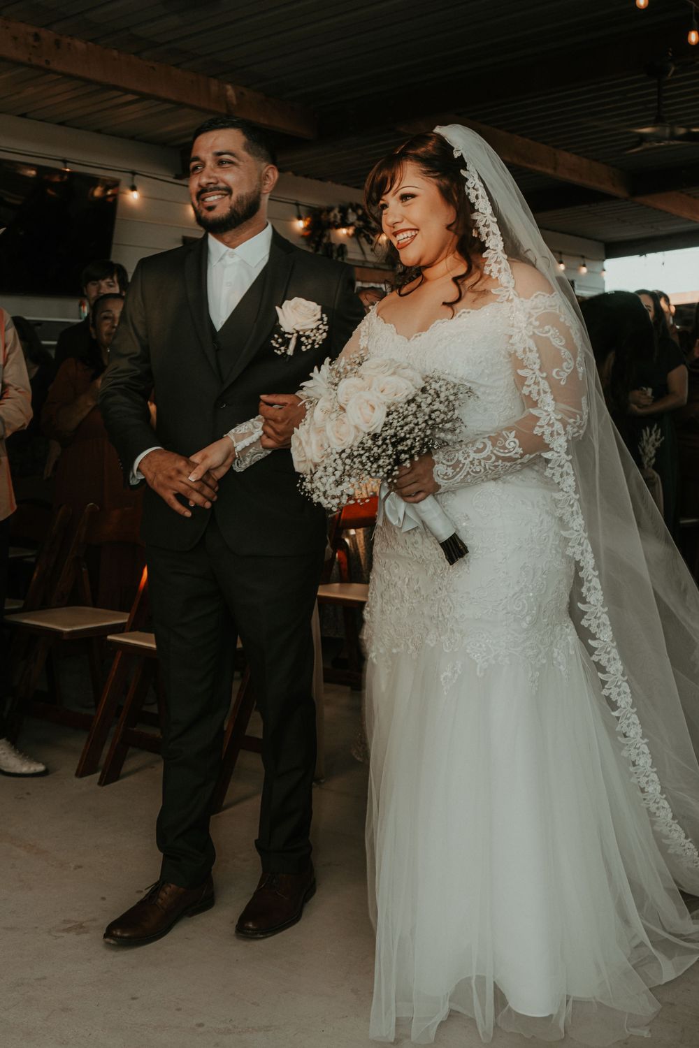 A smiling bride in a lace gown and veil holds a bouquet while walking the aisle with her groom in a dark suit.