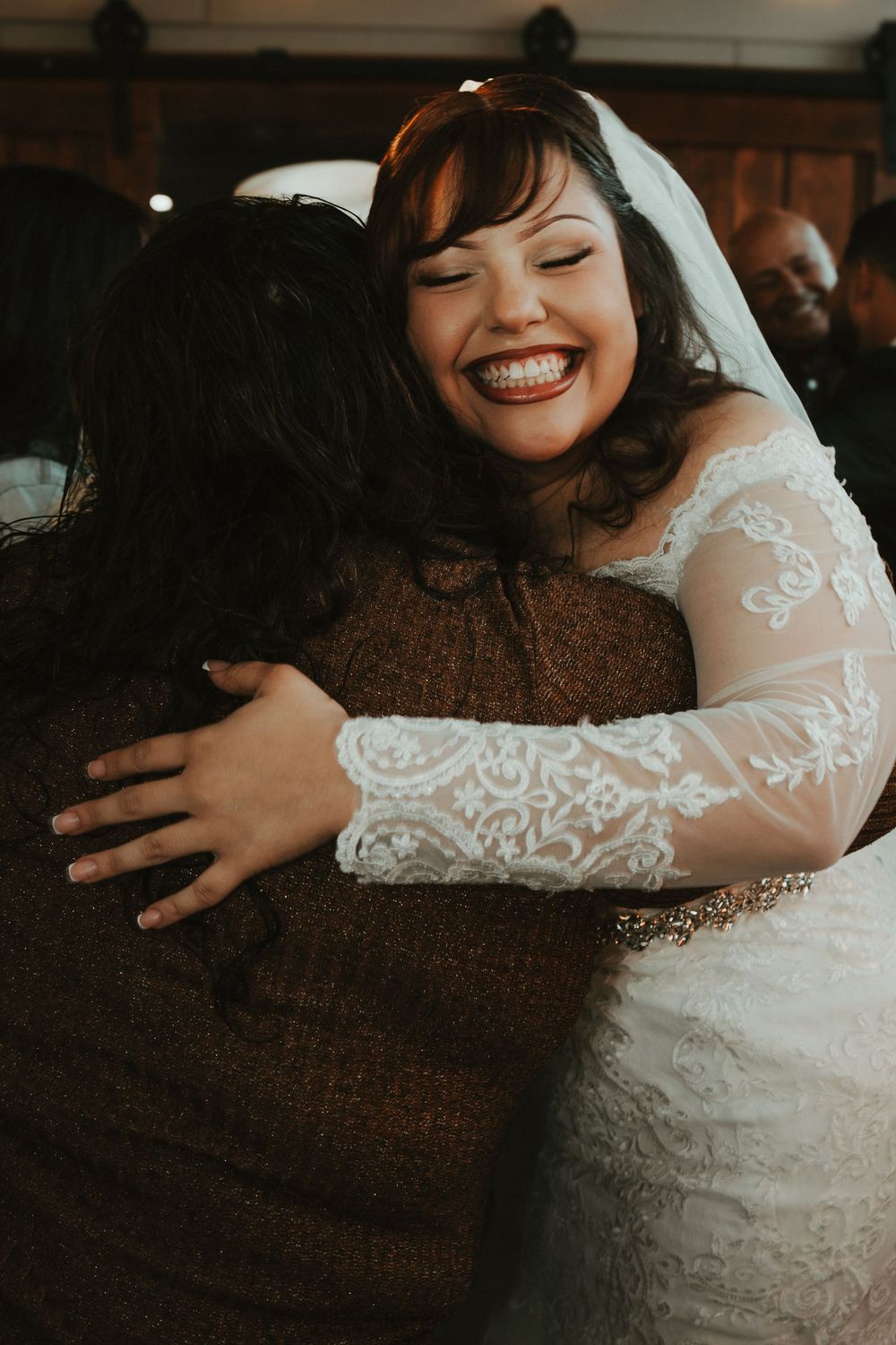 Smiling bride in lace wedding dress and veil hugging a guest at her wedding reception.