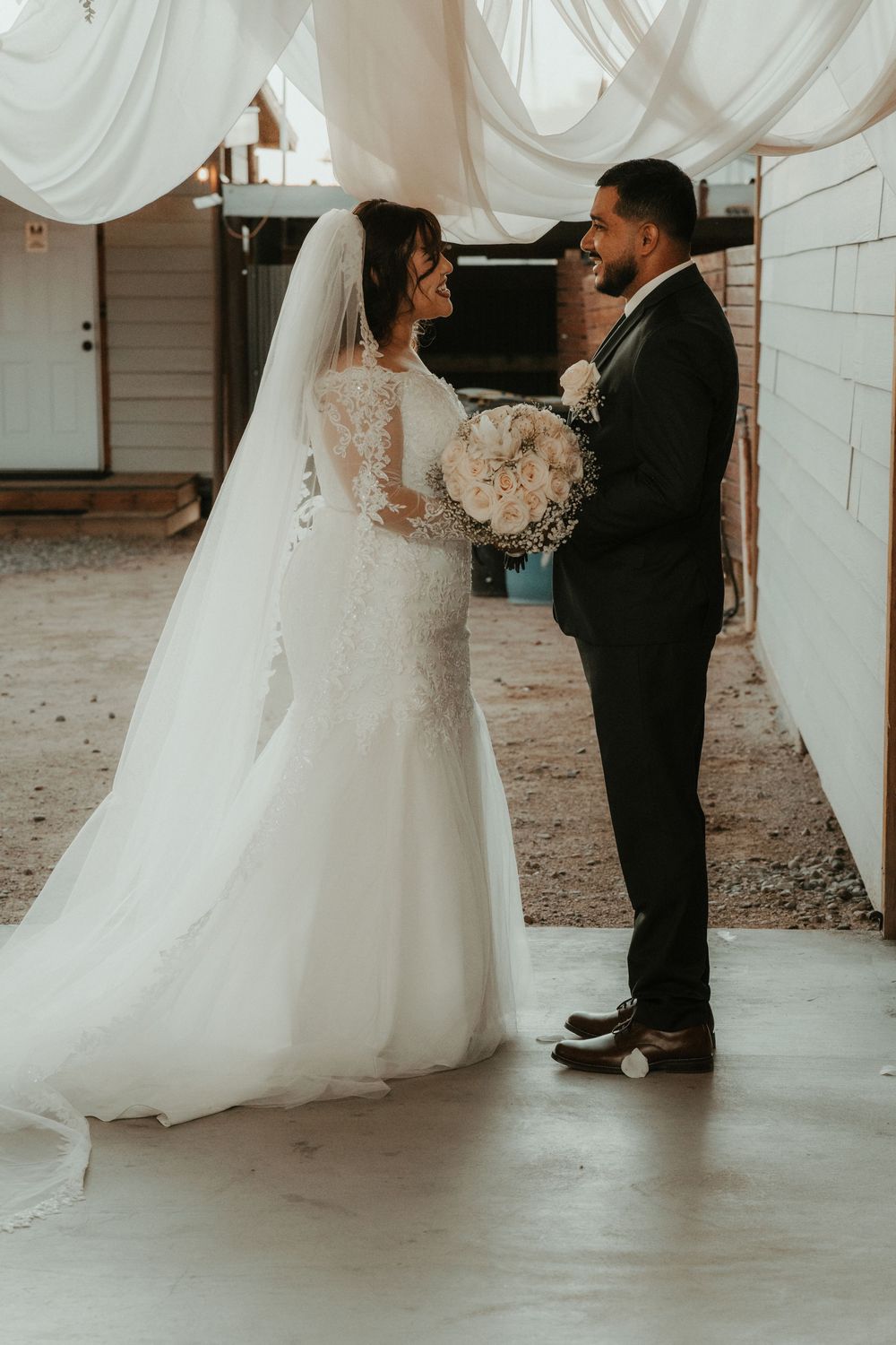 Bride in lace mermaid gown with veil holds bouquet facing groom in black suit during wedding ceremony.