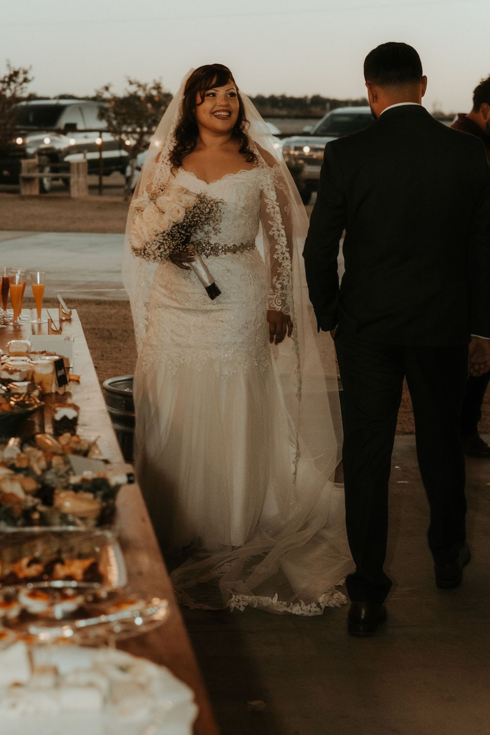 Bride in lace wedding gown and veil holds bouquet beside groom in black suit at outdoor wedding reception.