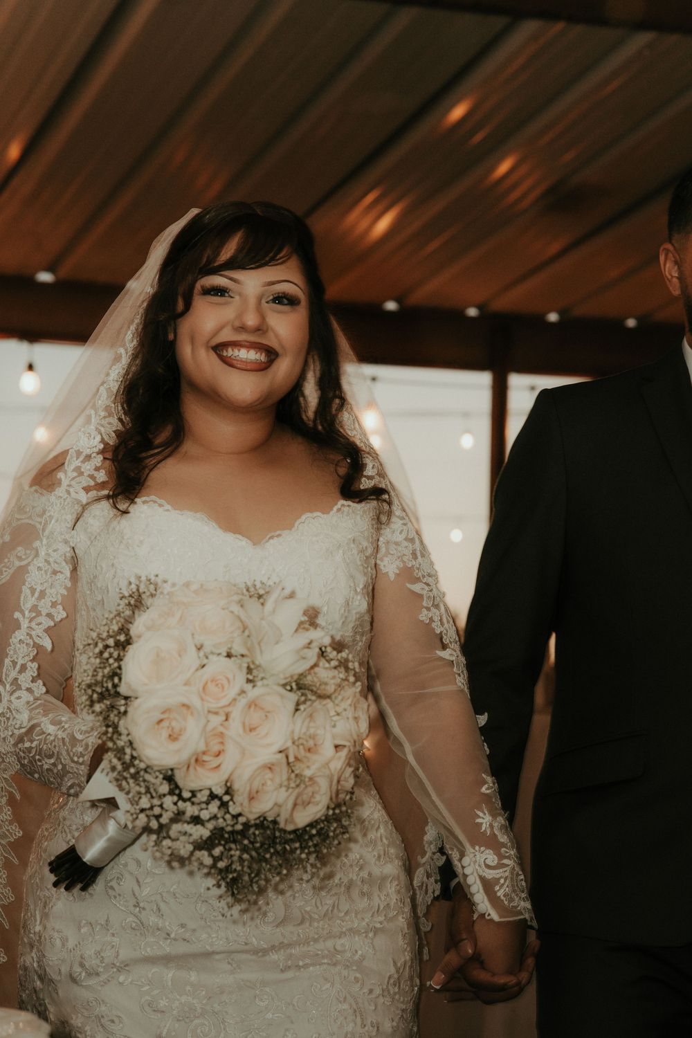 Smiling bride in lace long-sleeve wedding dress holding white rose bouquet, wearing veil, standing beside groom indoors.
