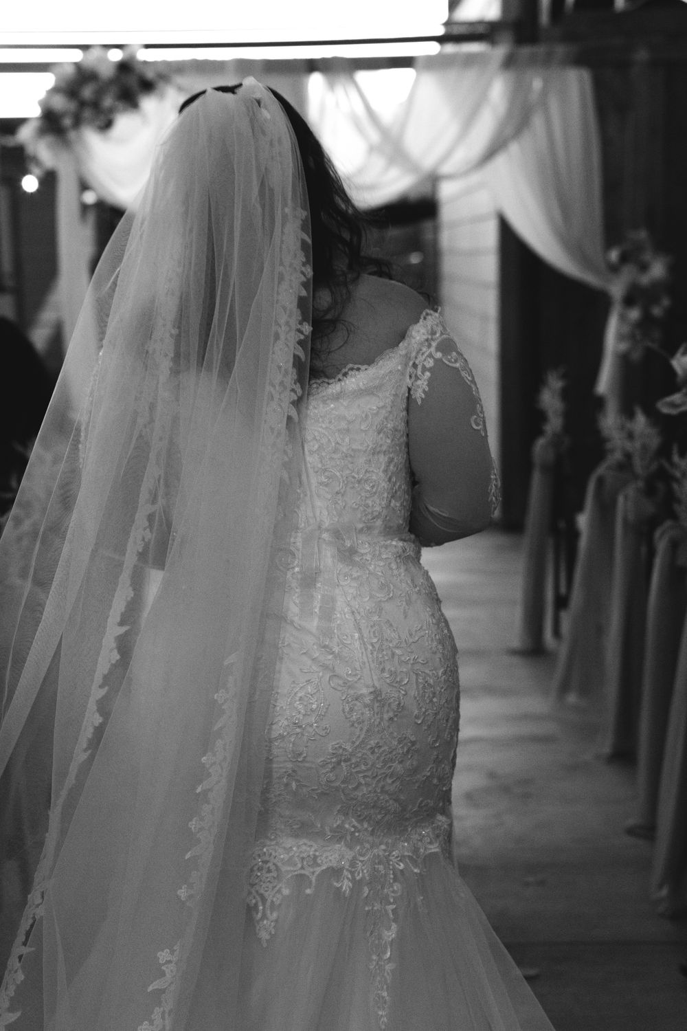 Black and white photo of a bride in a lace mermaid gown and long veil walking down the aisle.