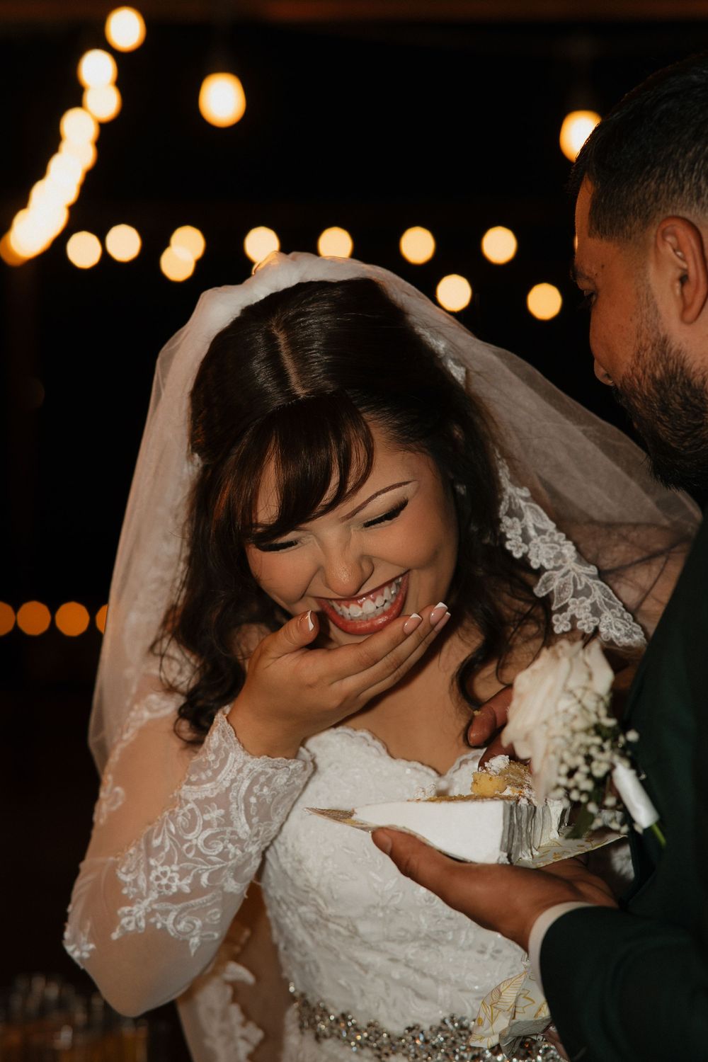 Joyful bride in lace wedding dress and veil laughs while eating cake at her wedding reception.