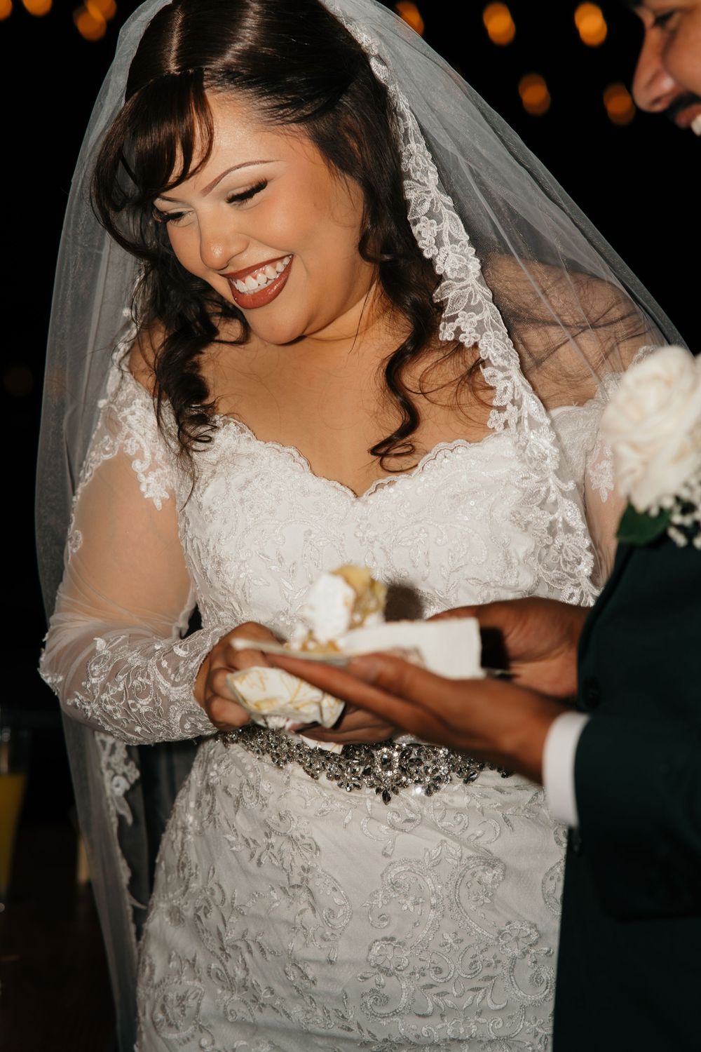 Smiling bride in lace gown and veil receives wedding cake from groom at evening reception.