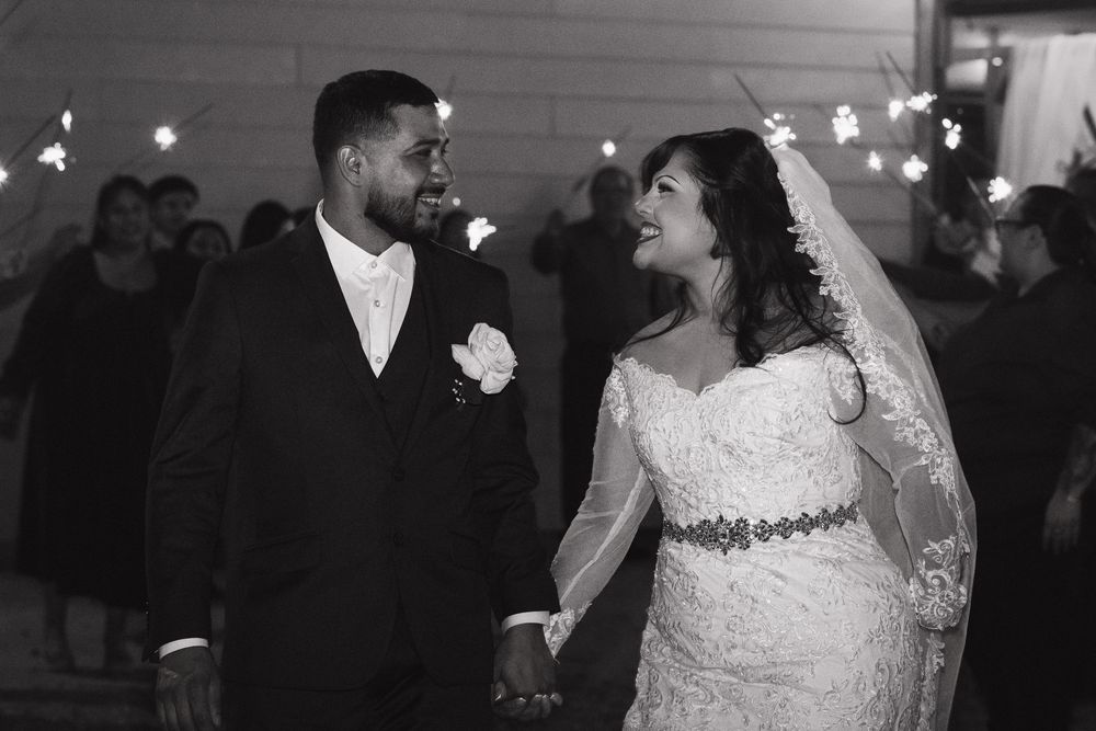 Black and white photo of a smiling bride and groom holding hands at their wedding reception with sparklers in background.