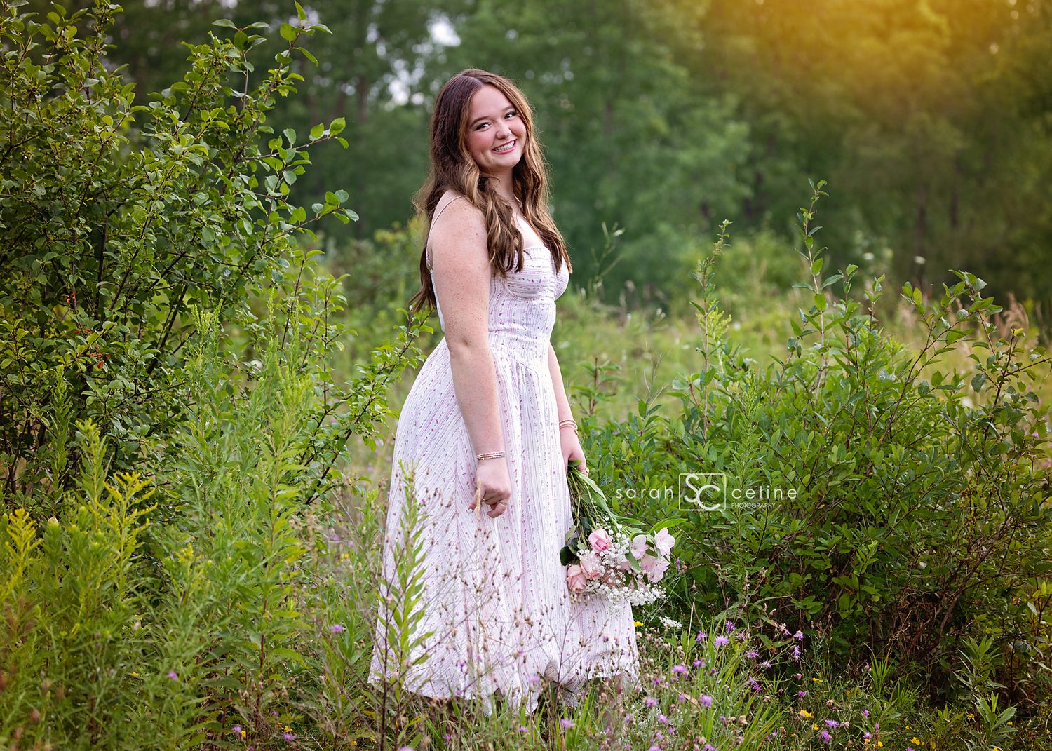 Young woman in white lace dress holding bouquet, smiling in lush green meadow during golden hour.