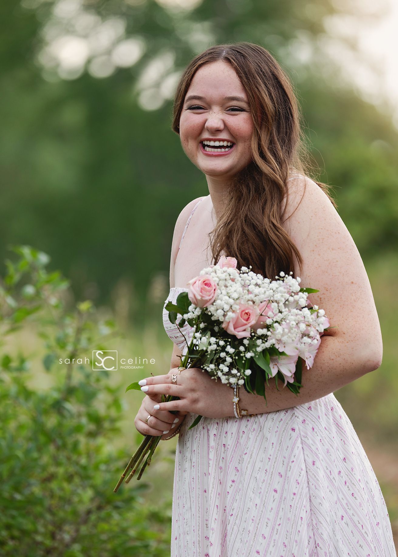 Smiling young woman in pink dress holding bouquet of pink roses and baby's breath outdoors.