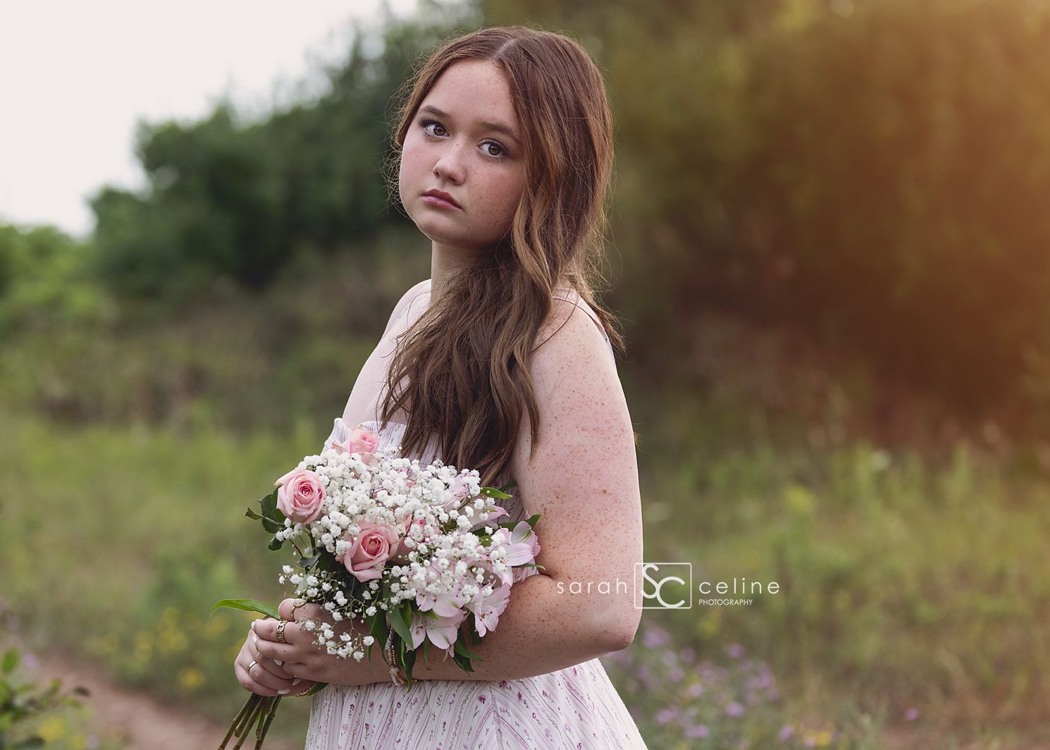 Young girl in white lace dress holding pink roses and baby's breath bouquet in a green field.