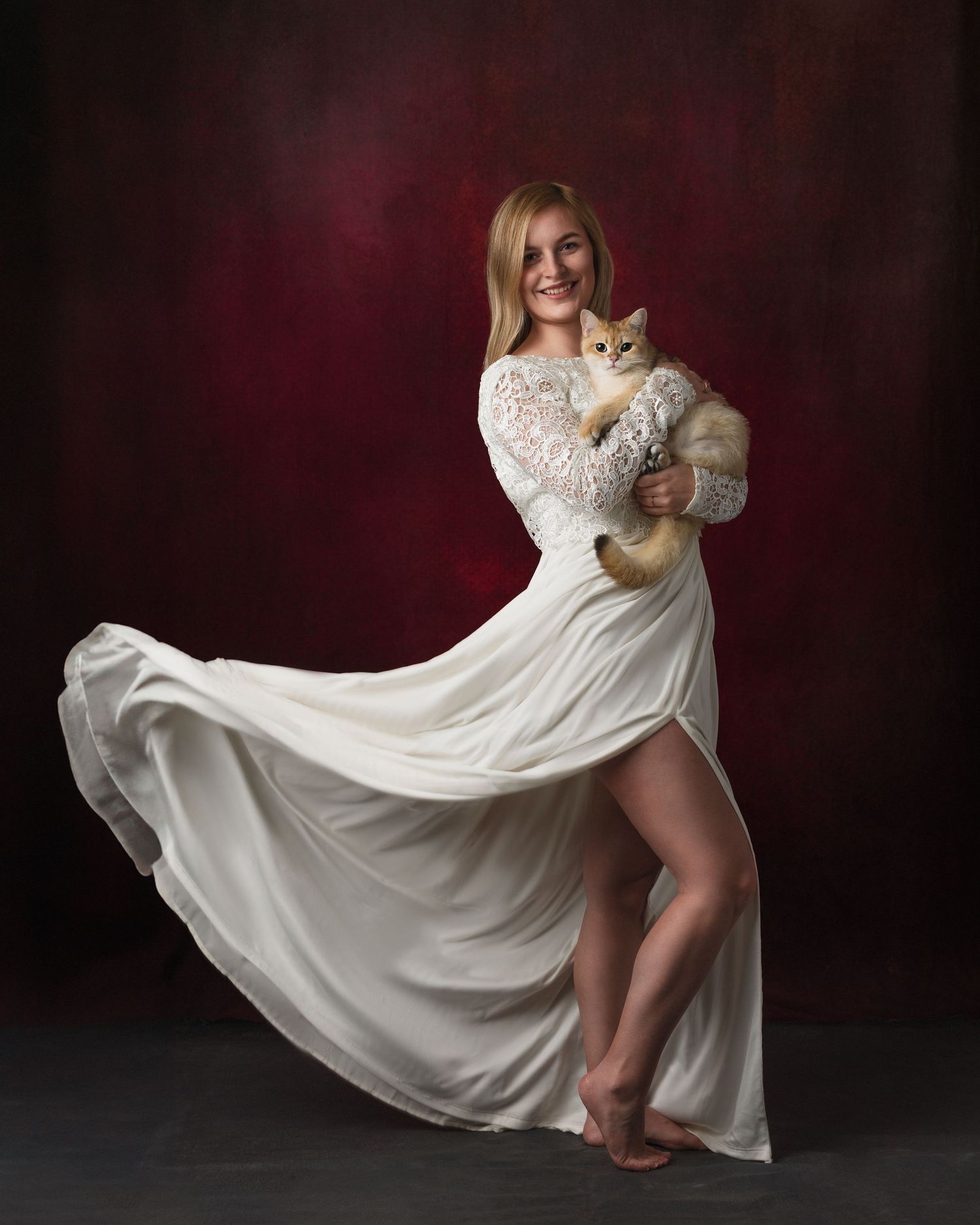 Blonde woman in flowing white lace gown holding an orange cat, posed against a dark red studio background.
